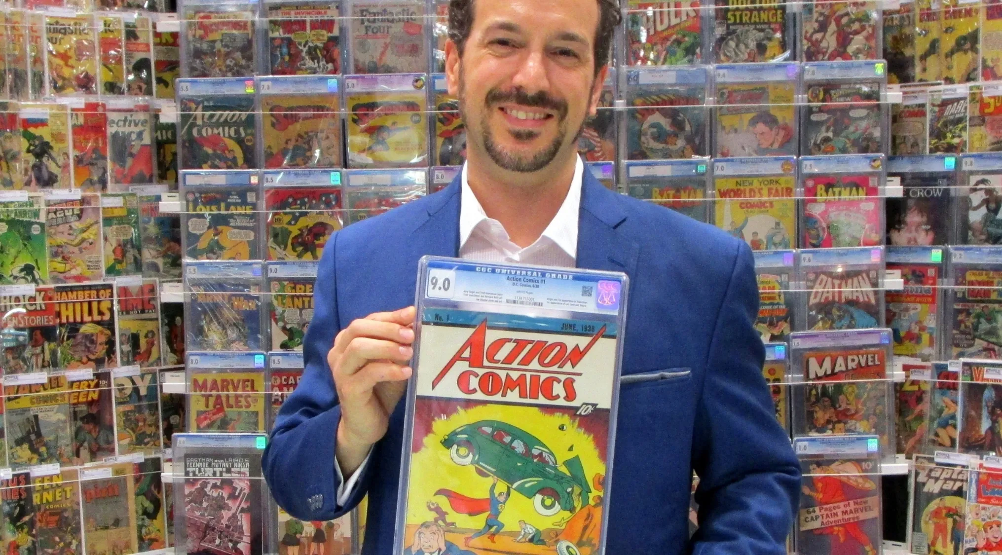 Man in blue suit holding a graded Action Comics No 1 comic surrounded by vintage comic books for price guessing in 2024.