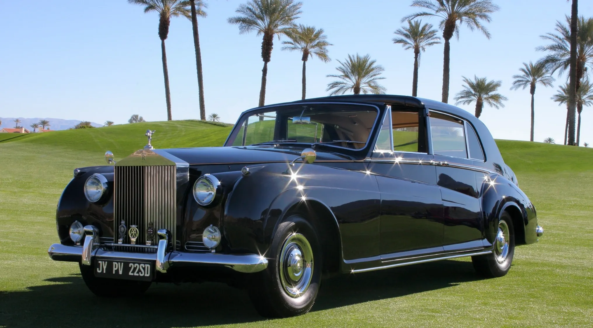 Classic black Phantom V Rolls Royce parked on green grass with palm trees in the background, shining in the sunlight.