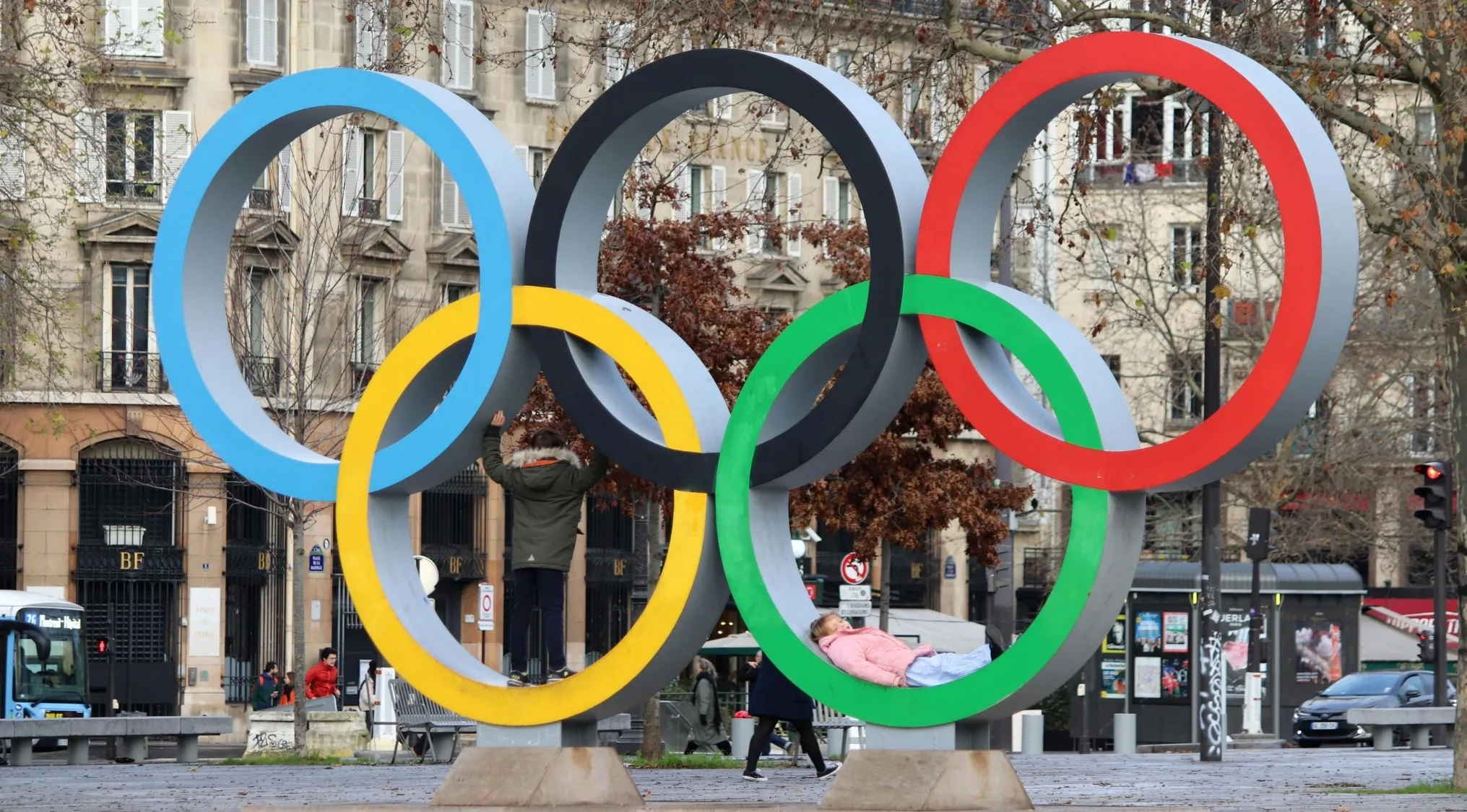 Large colorful Olympic rings sculpture in a city park with people interacting and buildings in the background.
