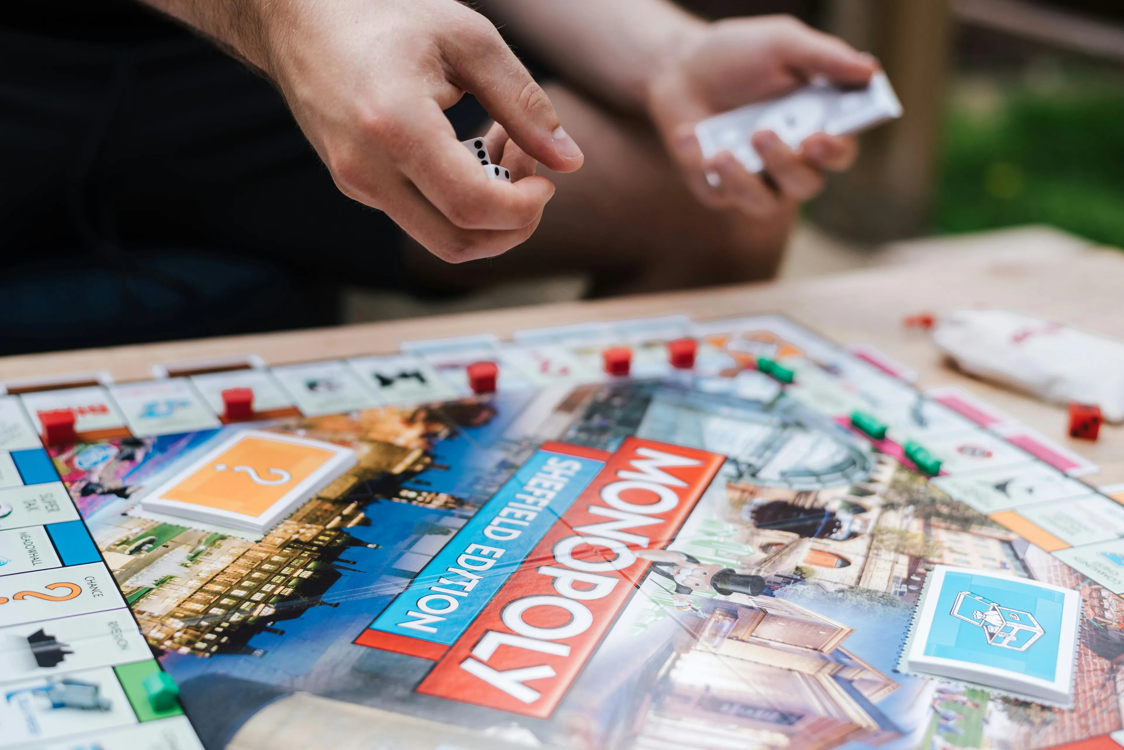 Close-up of hands playing Monopoly board game, focusing on dice and property cards during a game session outdoors.