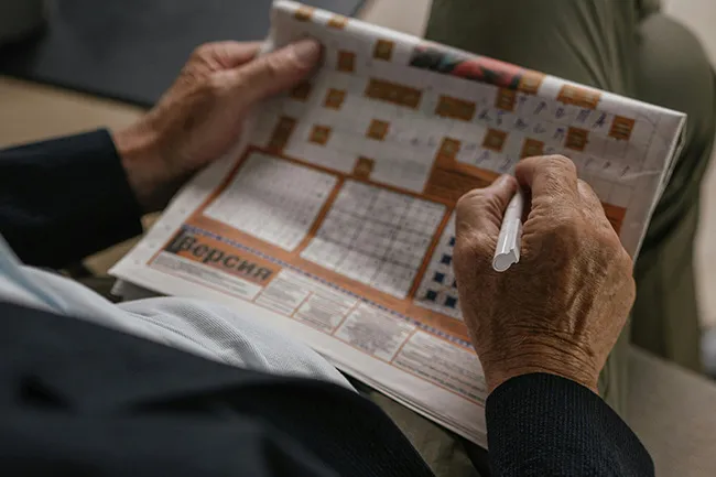 Elderly person solving a crossword puzzle with a pen, enjoying a quiet rainy-day activity indoors.