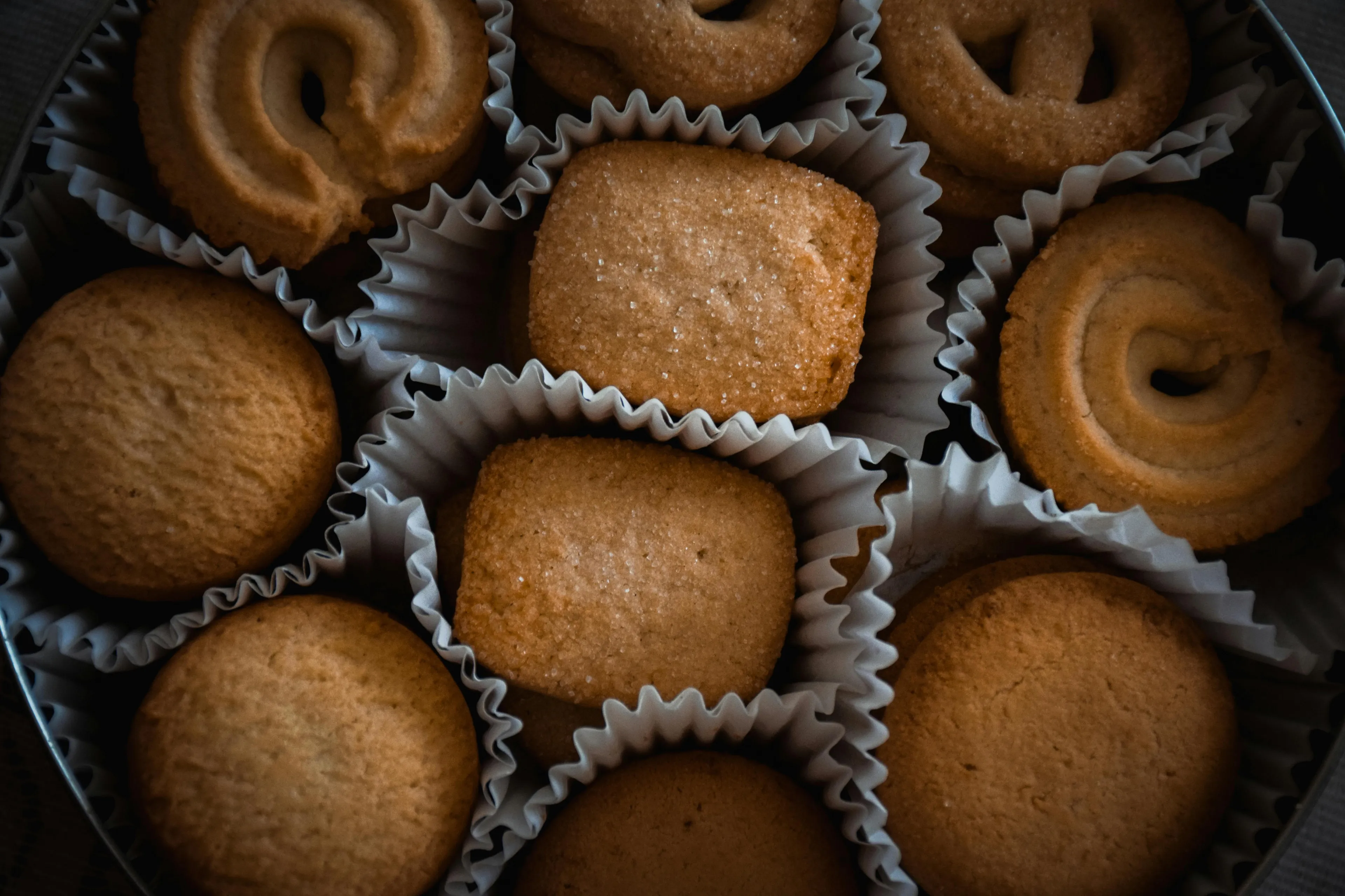 Assorted sugar cookies in paper cups arranged in a tin, perfect for a cozy rainy-day activity baking treat.