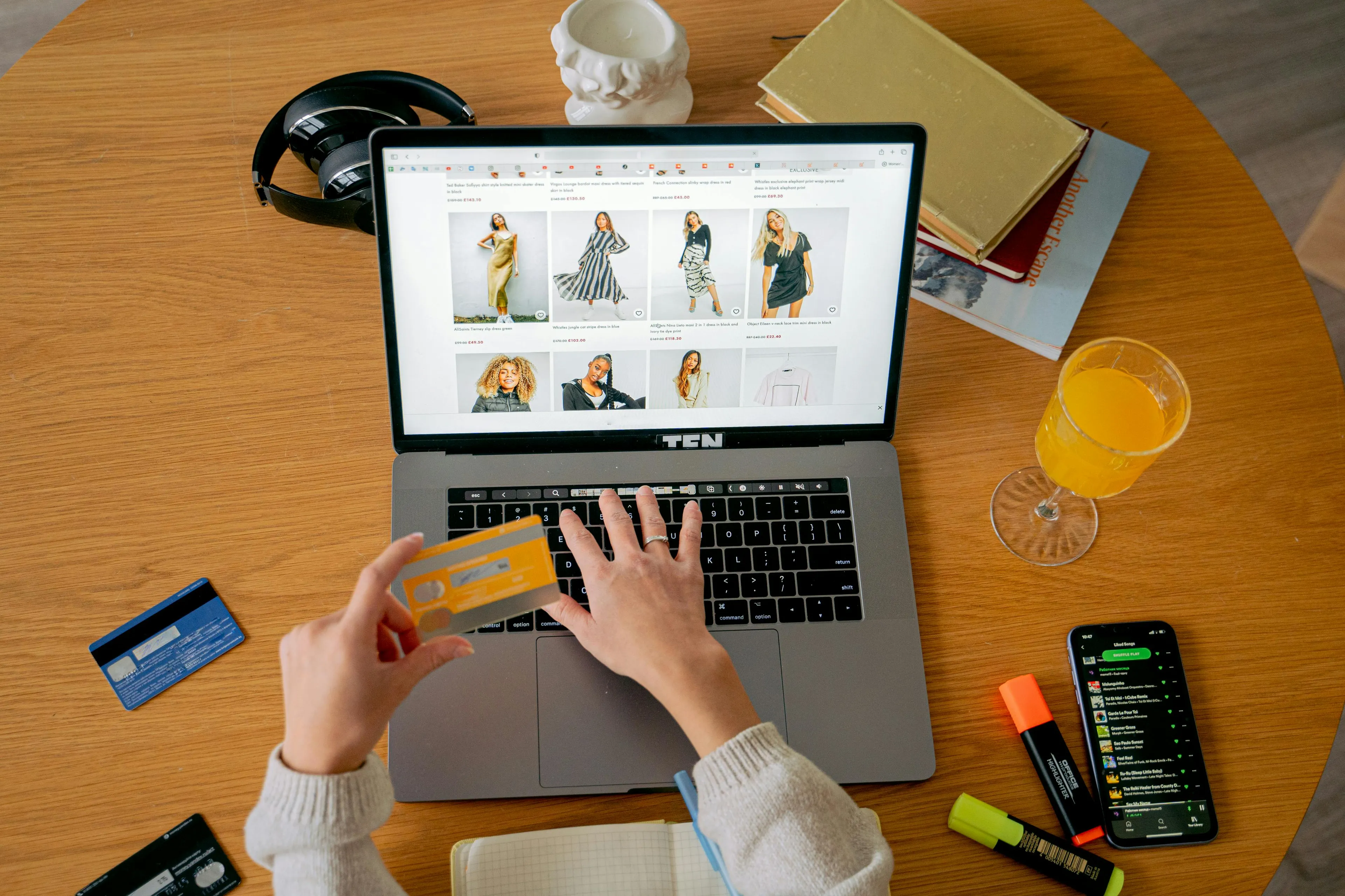 Person using a laptop for online shopping, holding a credit card with headphones, notebook, and drink nearby on table.