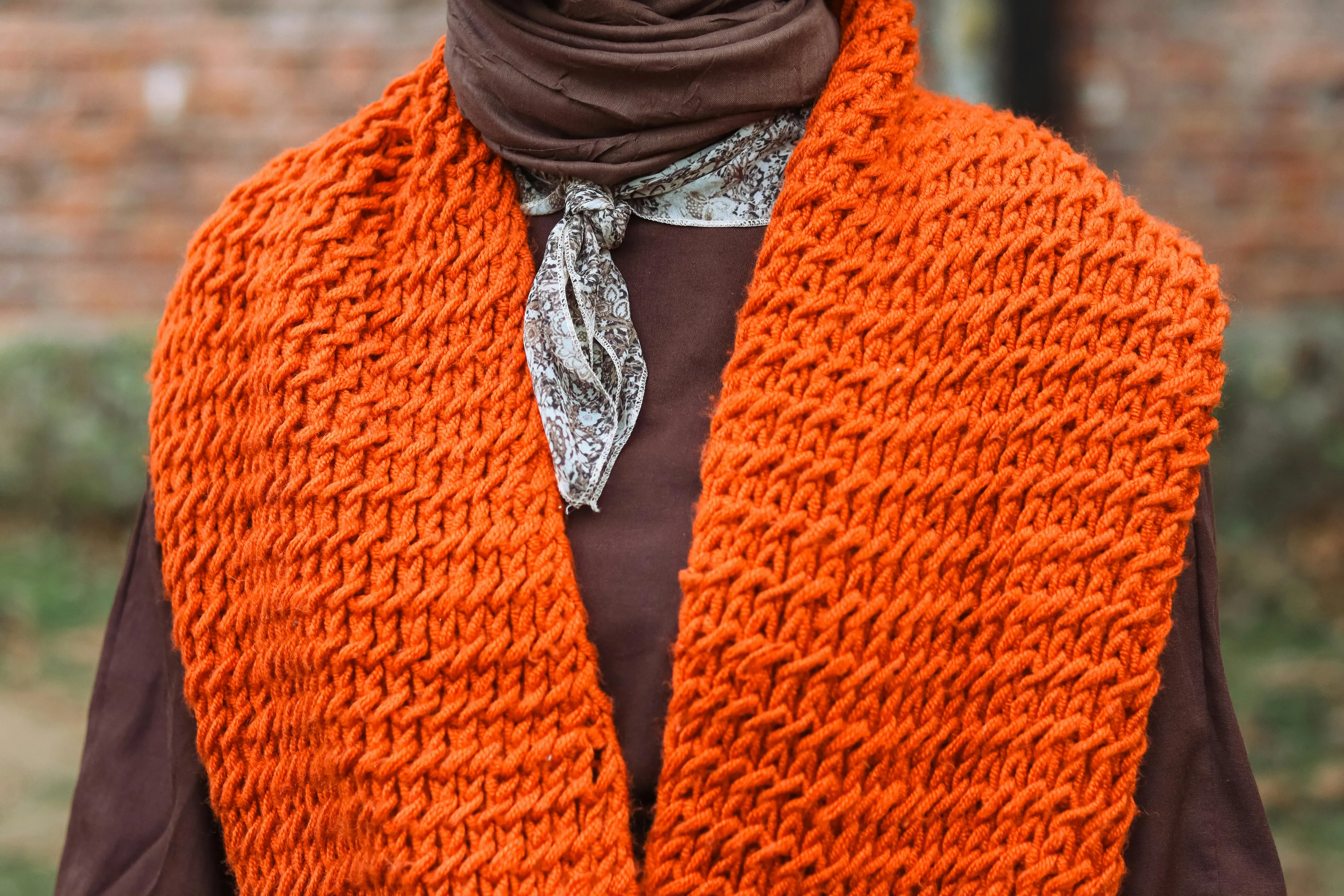 Handmade knitted scarf in bright orange worn over brown clothing, showcasing a detailed textured stitch pattern outdoors.