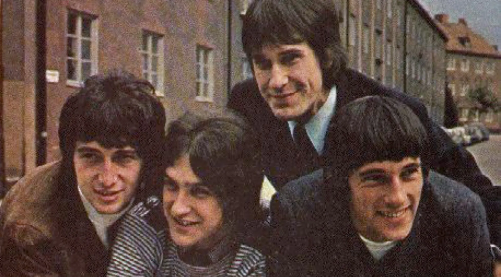The Kinks band members posing together outside on a street with brick buildings in the background