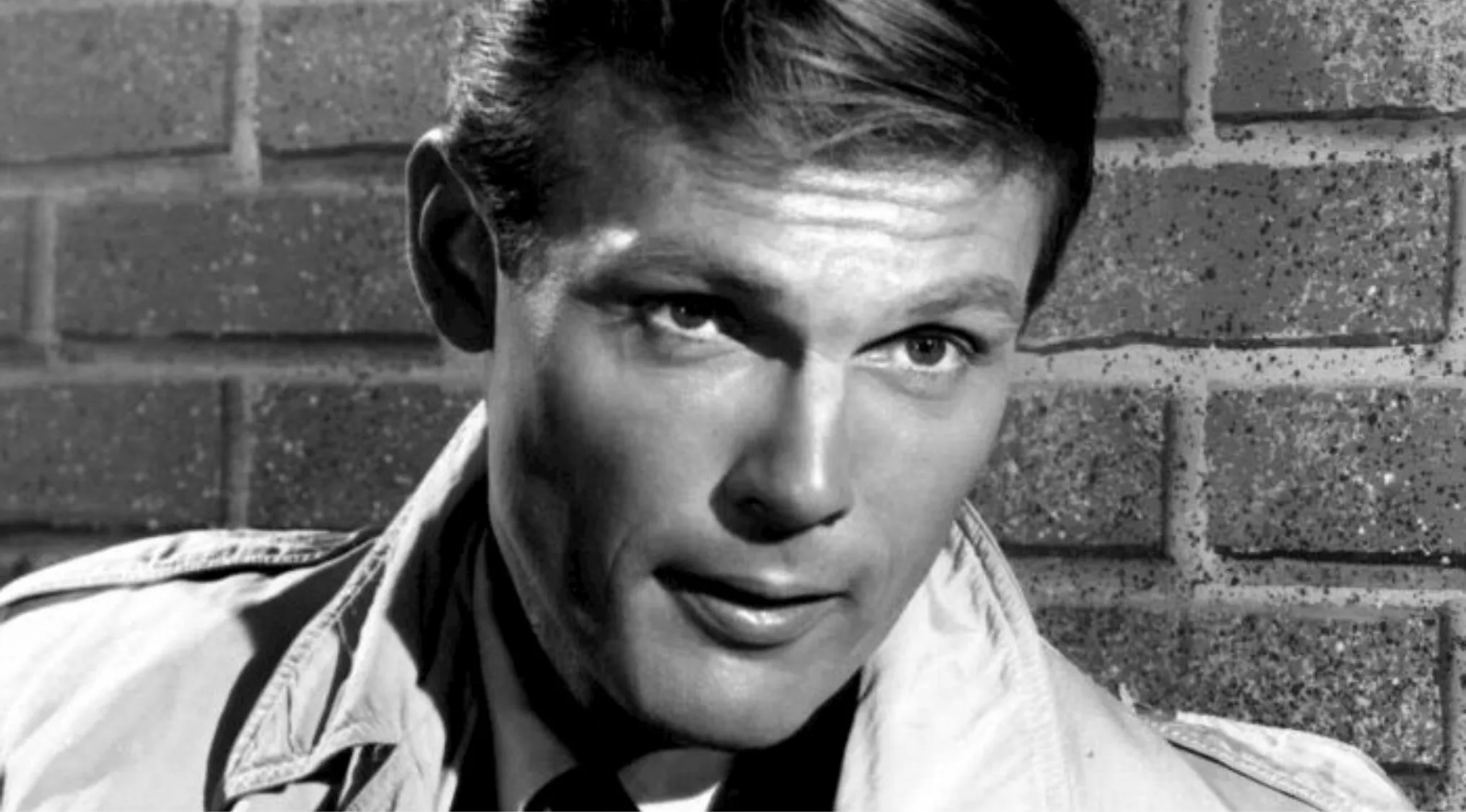 Black and white close-up of Adam West, star of the 60s superhero TV show, against a brick wall background