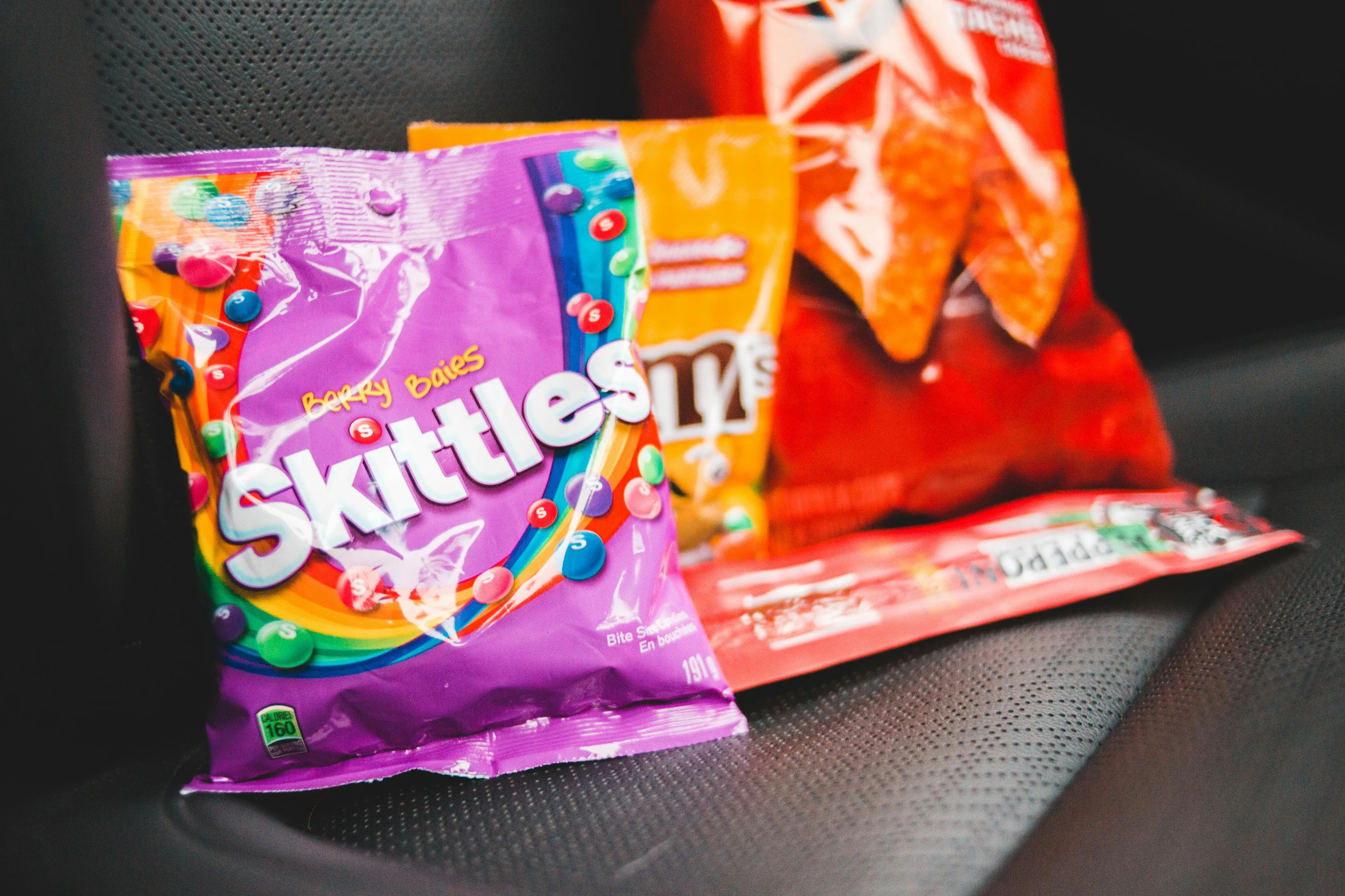 A bag of mystery snacks including colorful Skittles, M&M's, and Doritos arranged on a dark surface.
