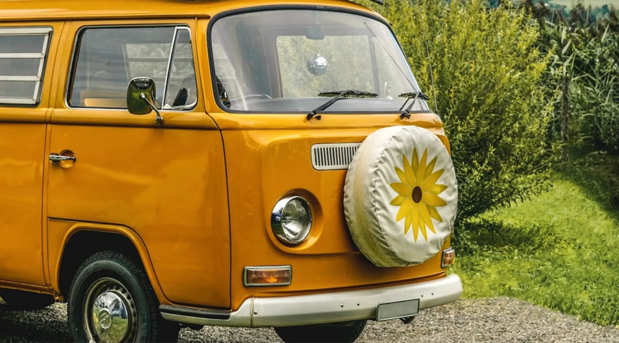Vintage orange camper van with a flower-covered spare tire parked on a gravel path surrounded by greenery.