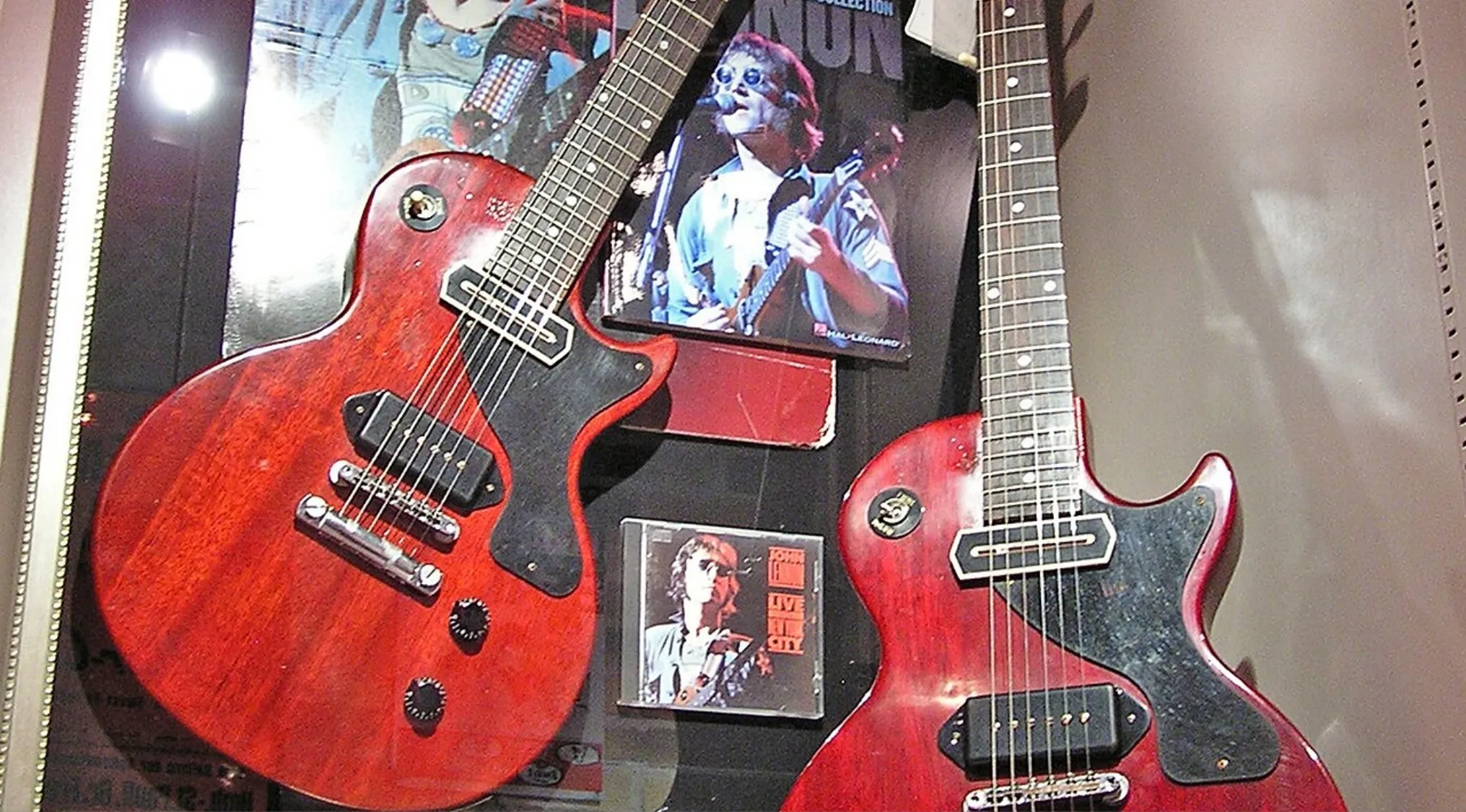 Two Gibson Les Paul Junior guitars displayed with a framed concert photo and album cover in the background.