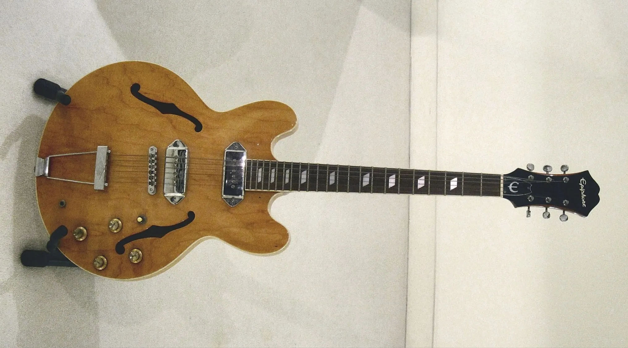 Natural wood finish Epiphone Casino hollow-body electric guitar displayed on a stand against a white wall.