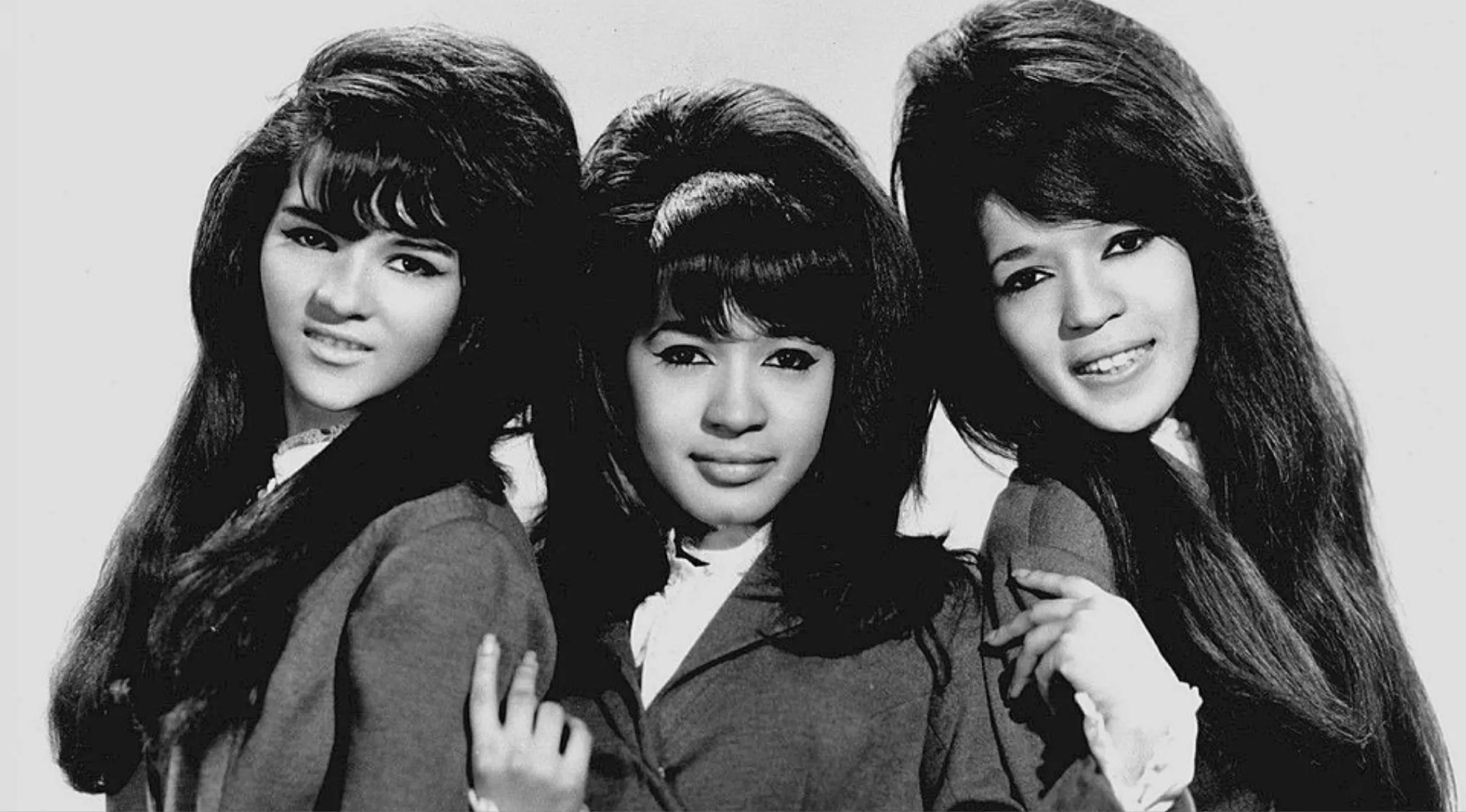 Three women with voluminous bouffant hairstyles wearing vintage-style coats posing together in a black and white photo.
