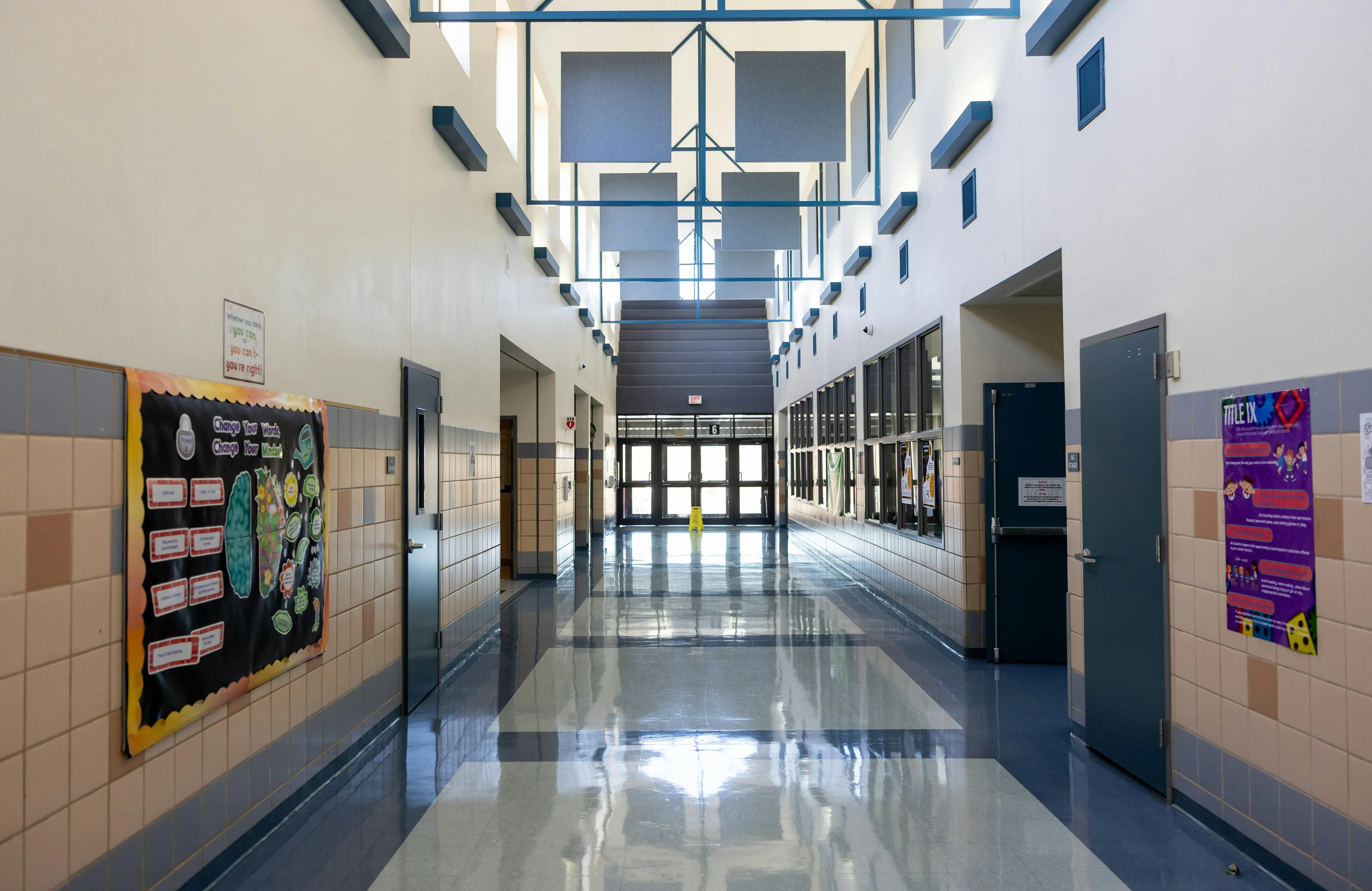 Bright school hallway with tiled walls, bulletin boards, and natural light shining through upper windows.