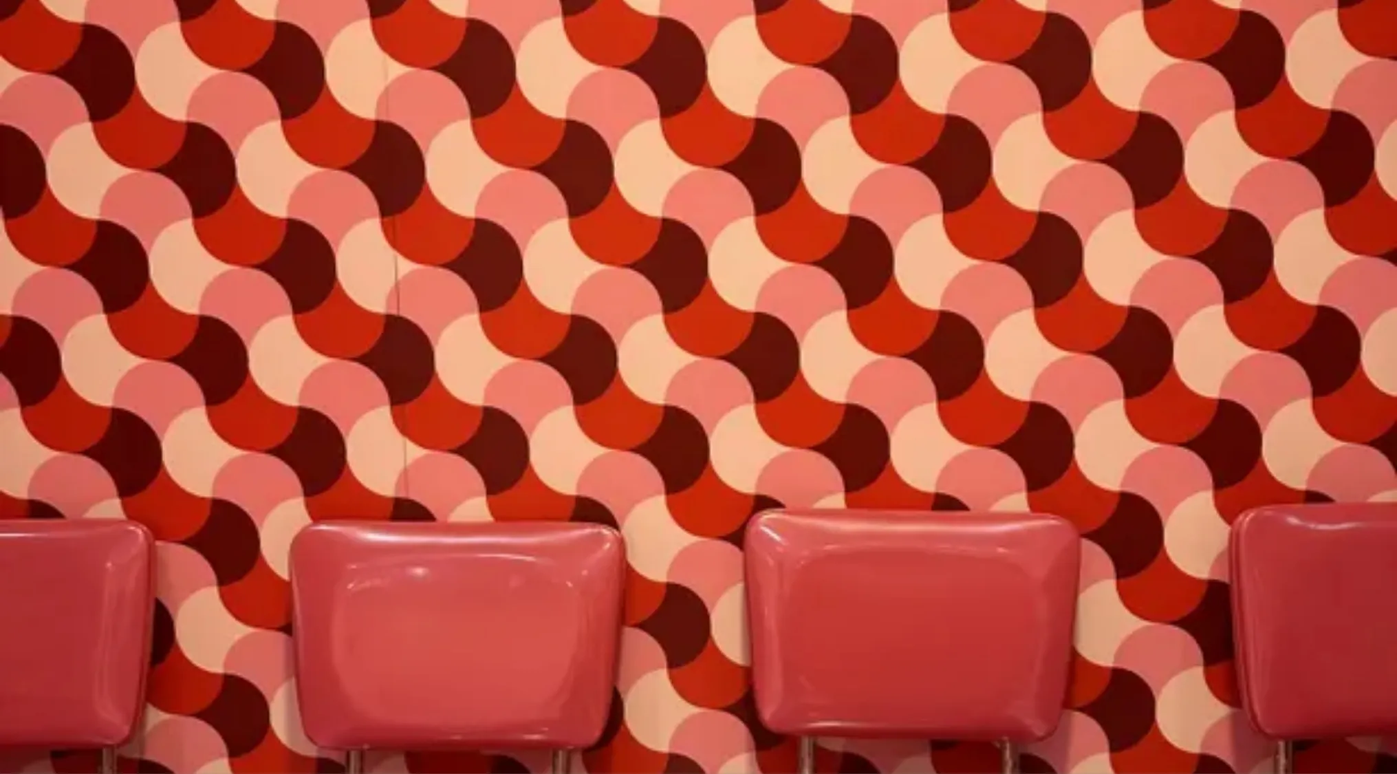 Four retro pink chairs against a wall with a vibrant geometric shapes pattern in red and pink tones.