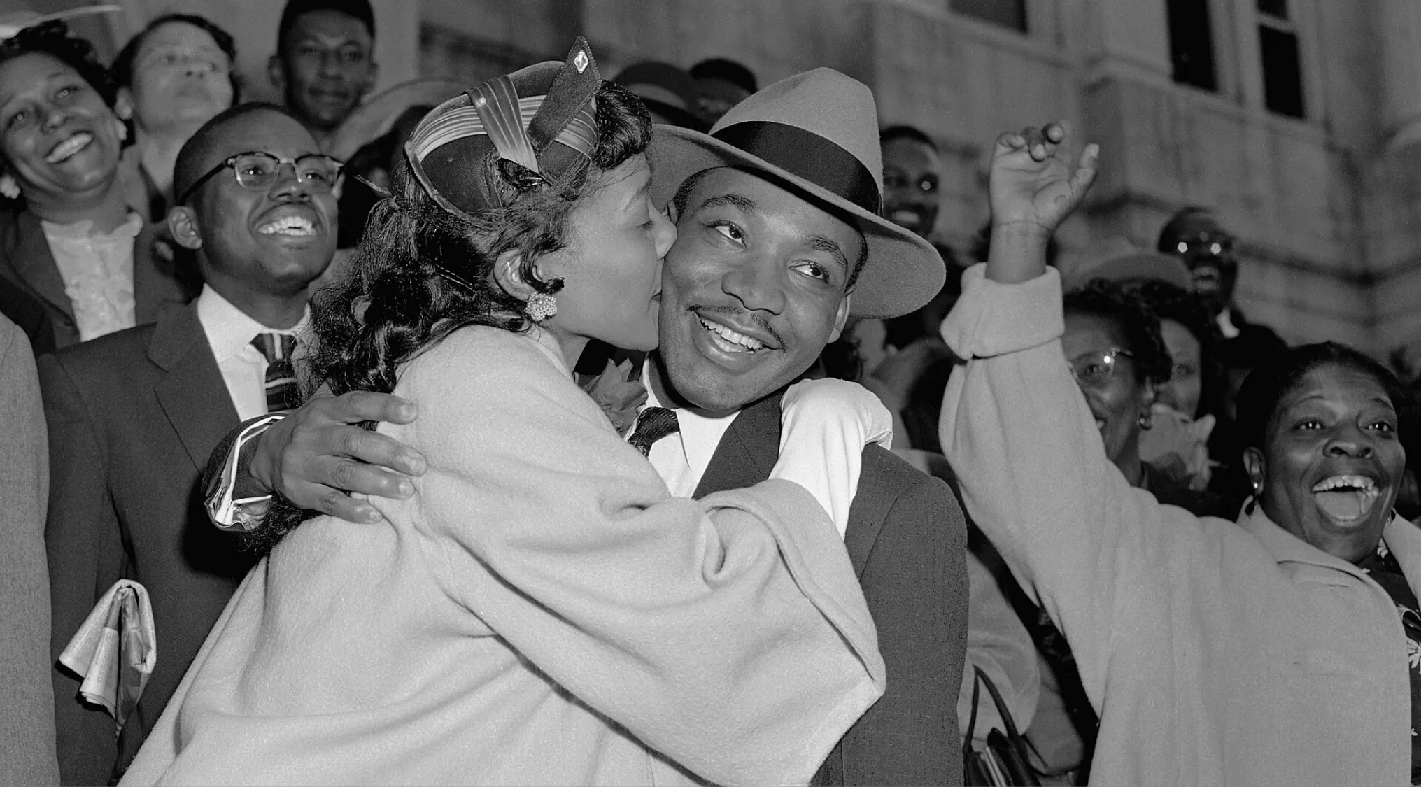 Martin Luther King Jr. smiling while being hugged and kissed by a woman surrounded by a cheering crowd.