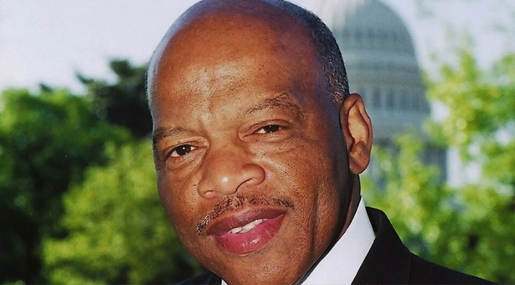 Close-up of civil rights leader John Lewis smiling outdoors with the U.S. Capitol blurred in the background.