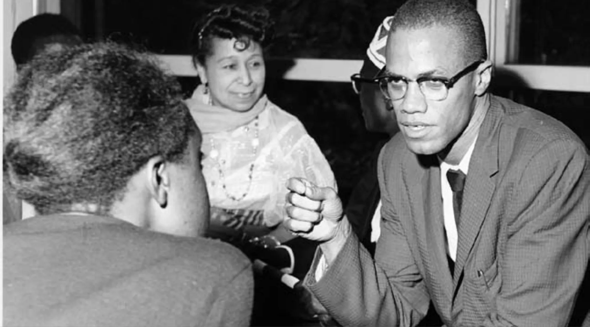 Malcolm X speaking and gesturing seriously with a group of people in a historical black and white photo.