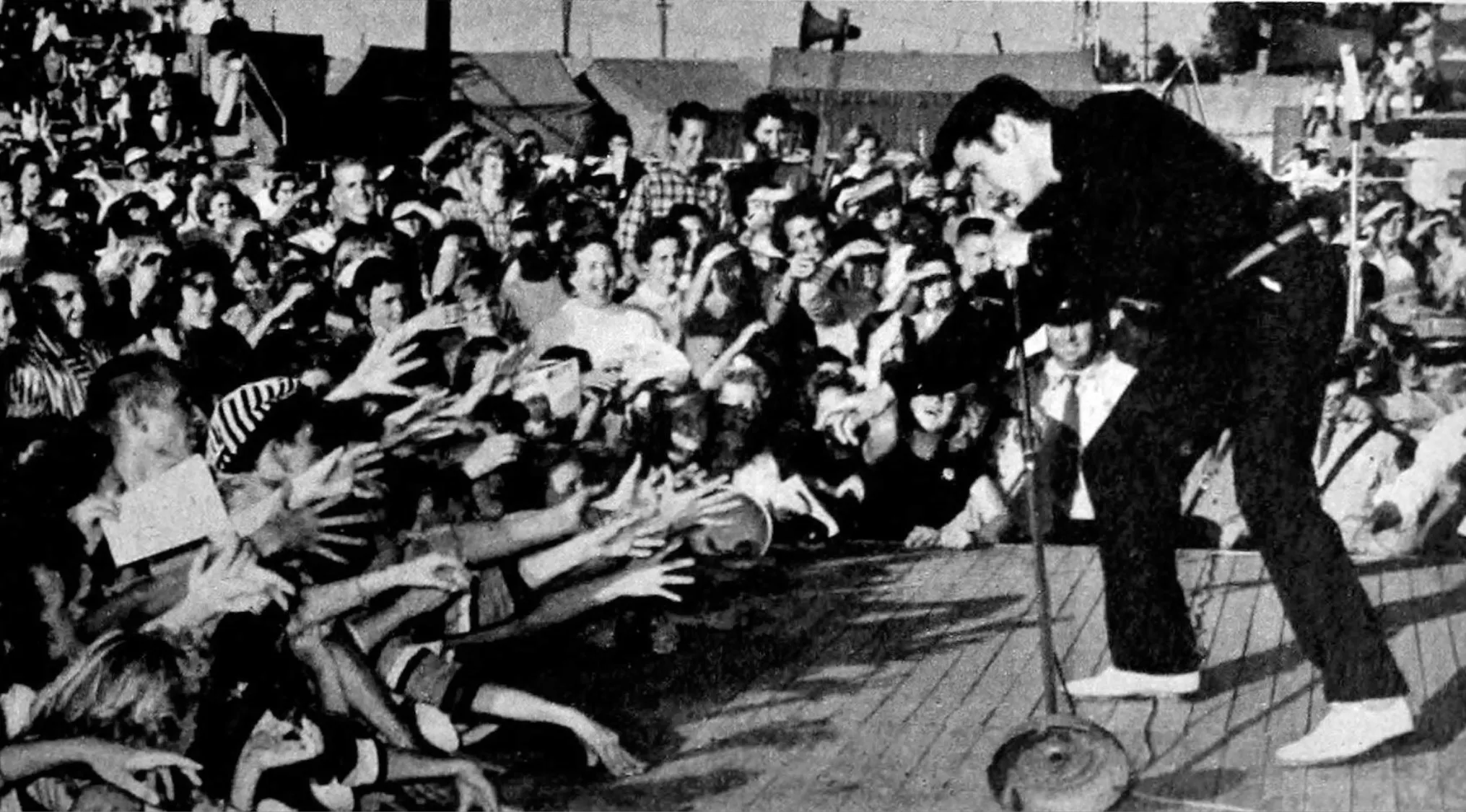 Elvis Presley performing on stage in the 60s, reaching out to excited fans at a live rock n roll concert.