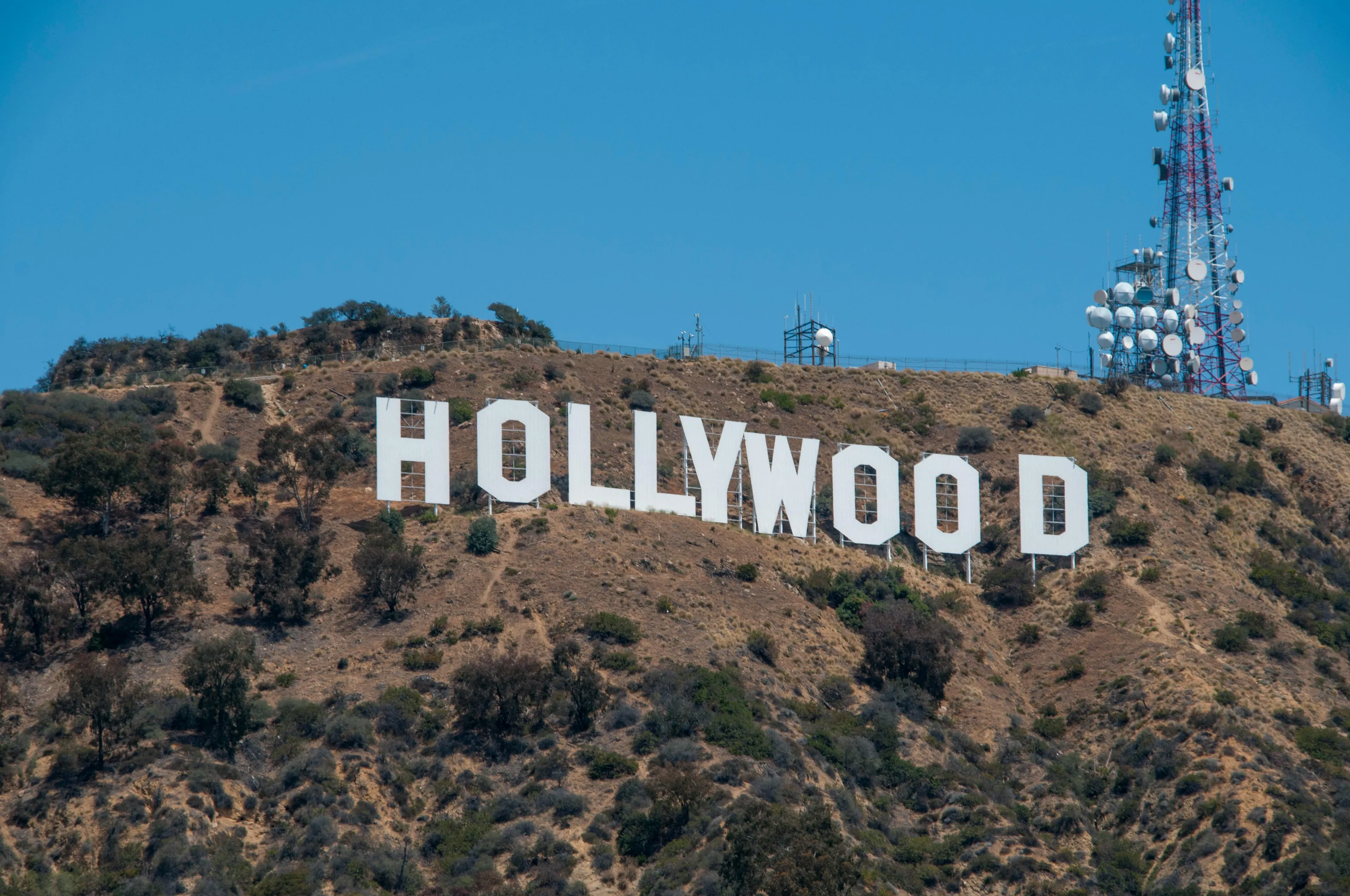 The Hollywood Sign on a dry hillside under a clear blue sky with communication towers nearby.