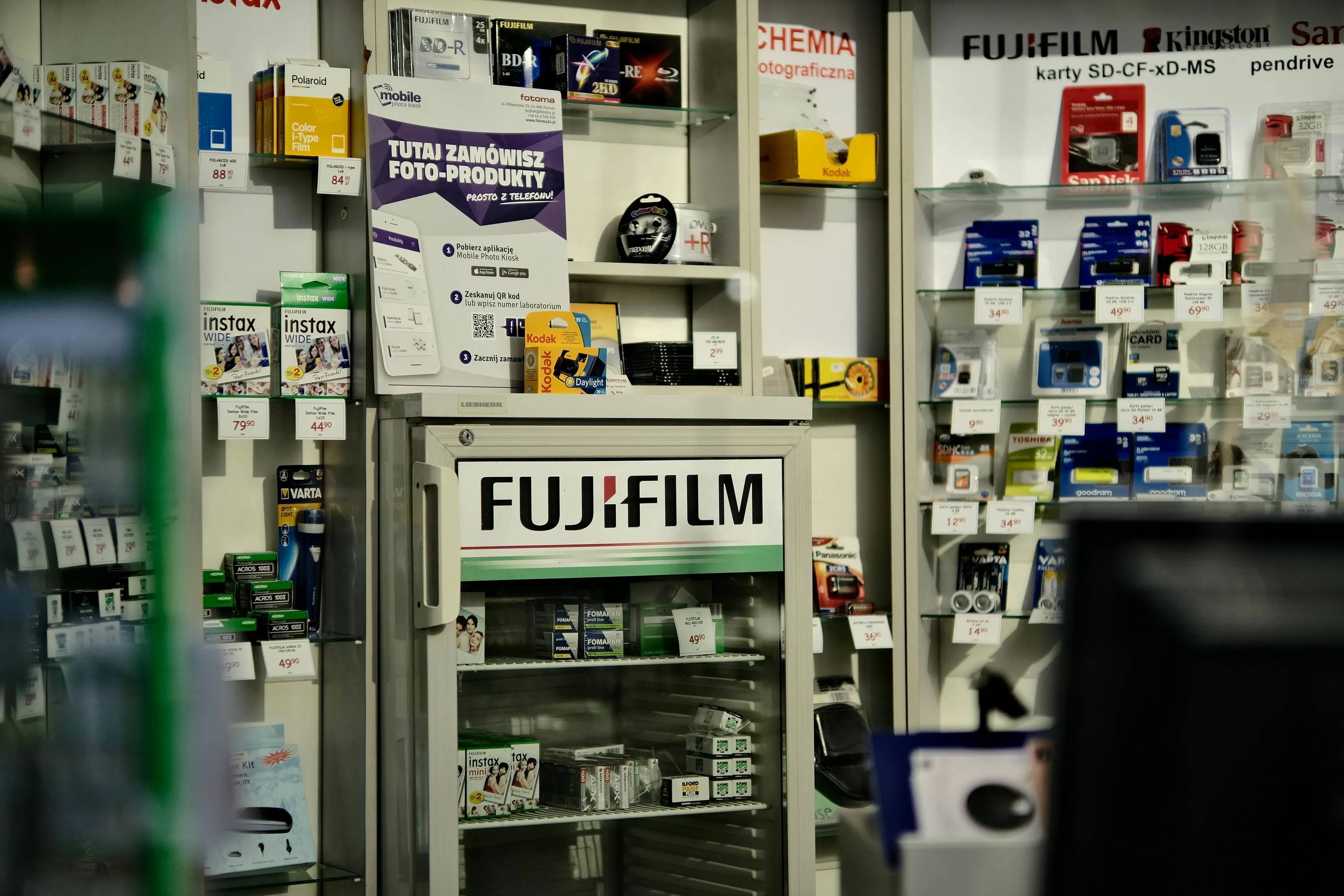 Electronics shop interior showing Fujifilm products and memory cards in a well-stocked neighborhood store display.