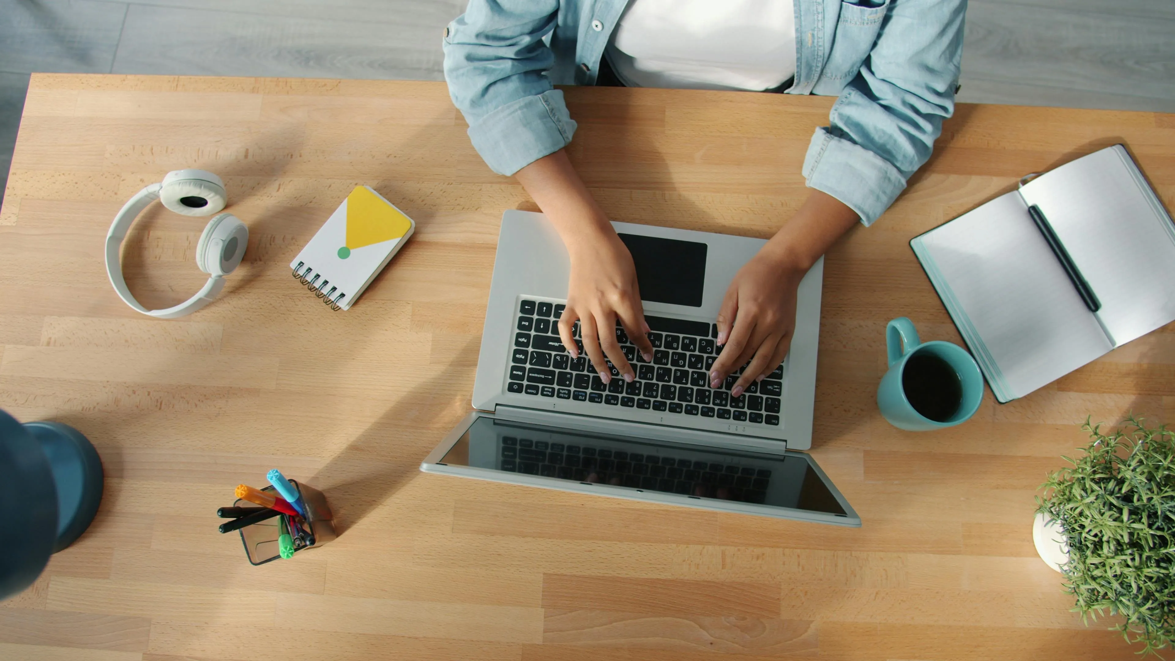 Person typing on laptop at work desk with headphones, notebook, coffee cup, and plant near workspace objects