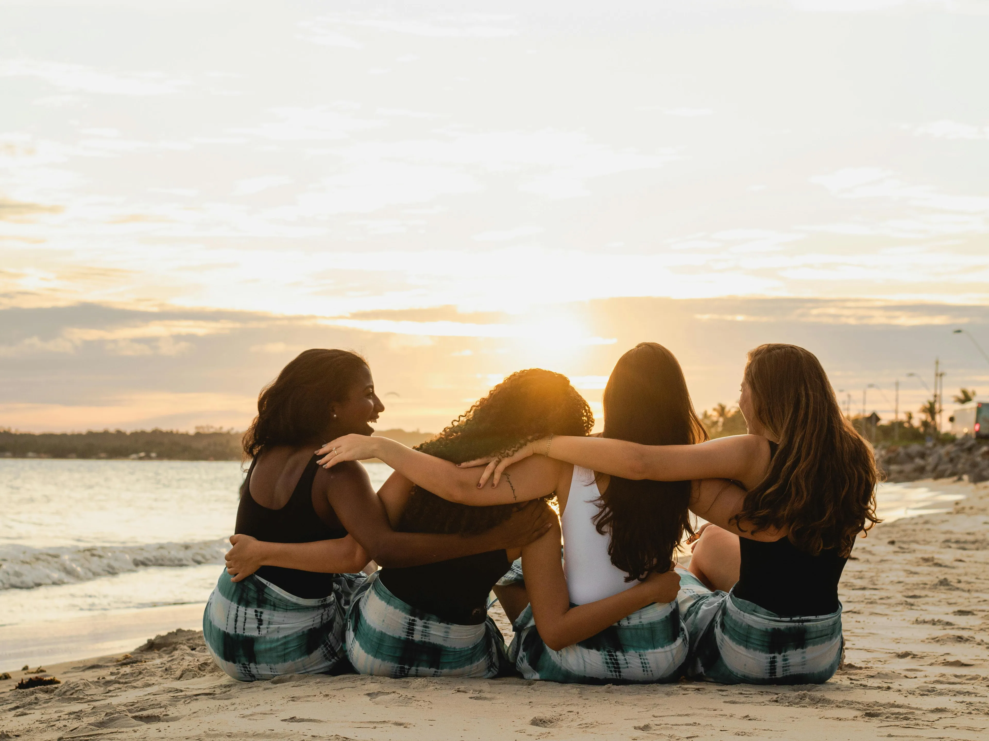 Four friends sitting on the beach at sunset with arms around each other, reflecting on how friends describe them.
