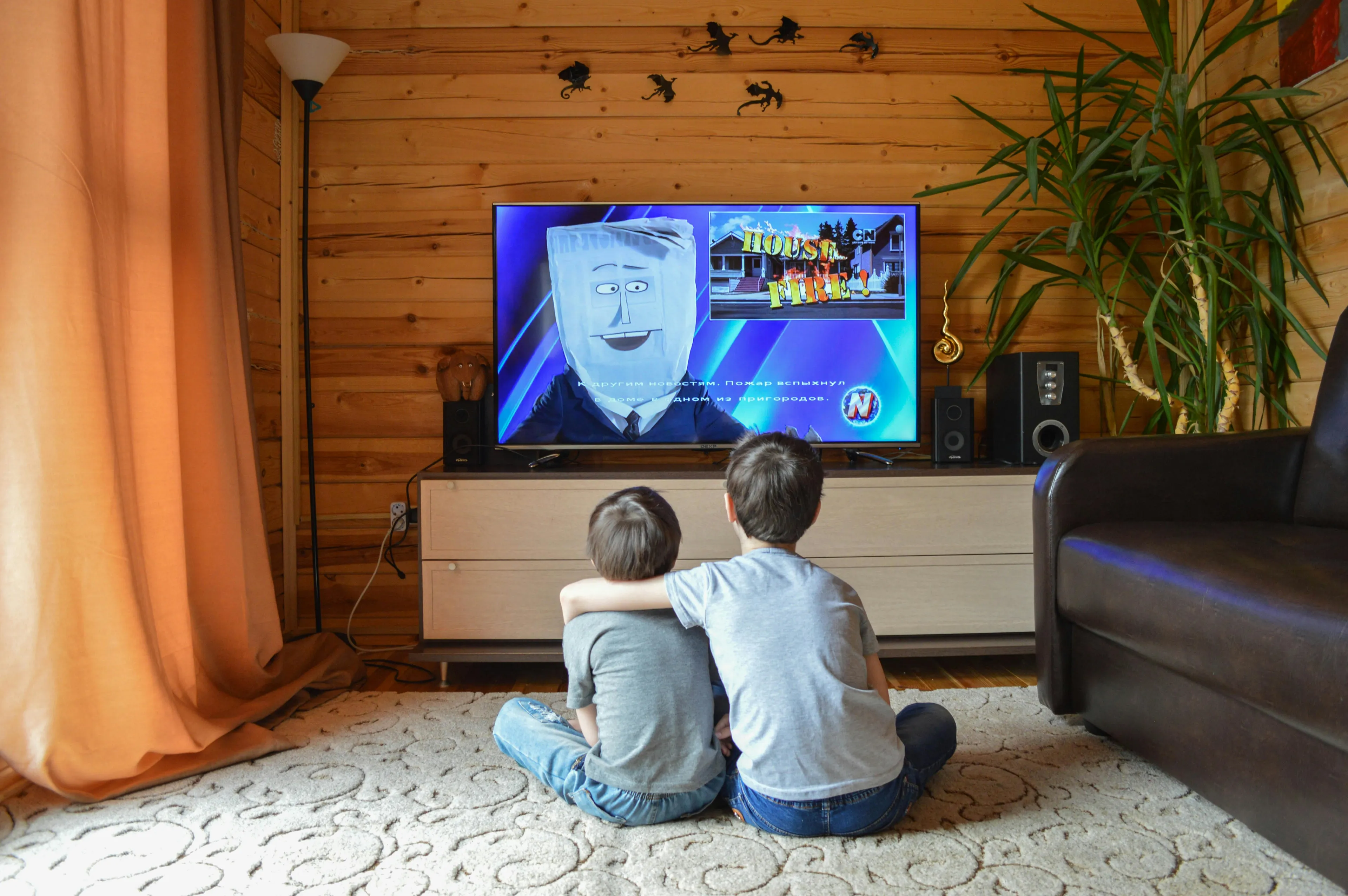 Two children sitting on a carpet watching a nostalgic childhood TV channel on a flat screen in a cozy wooden room.