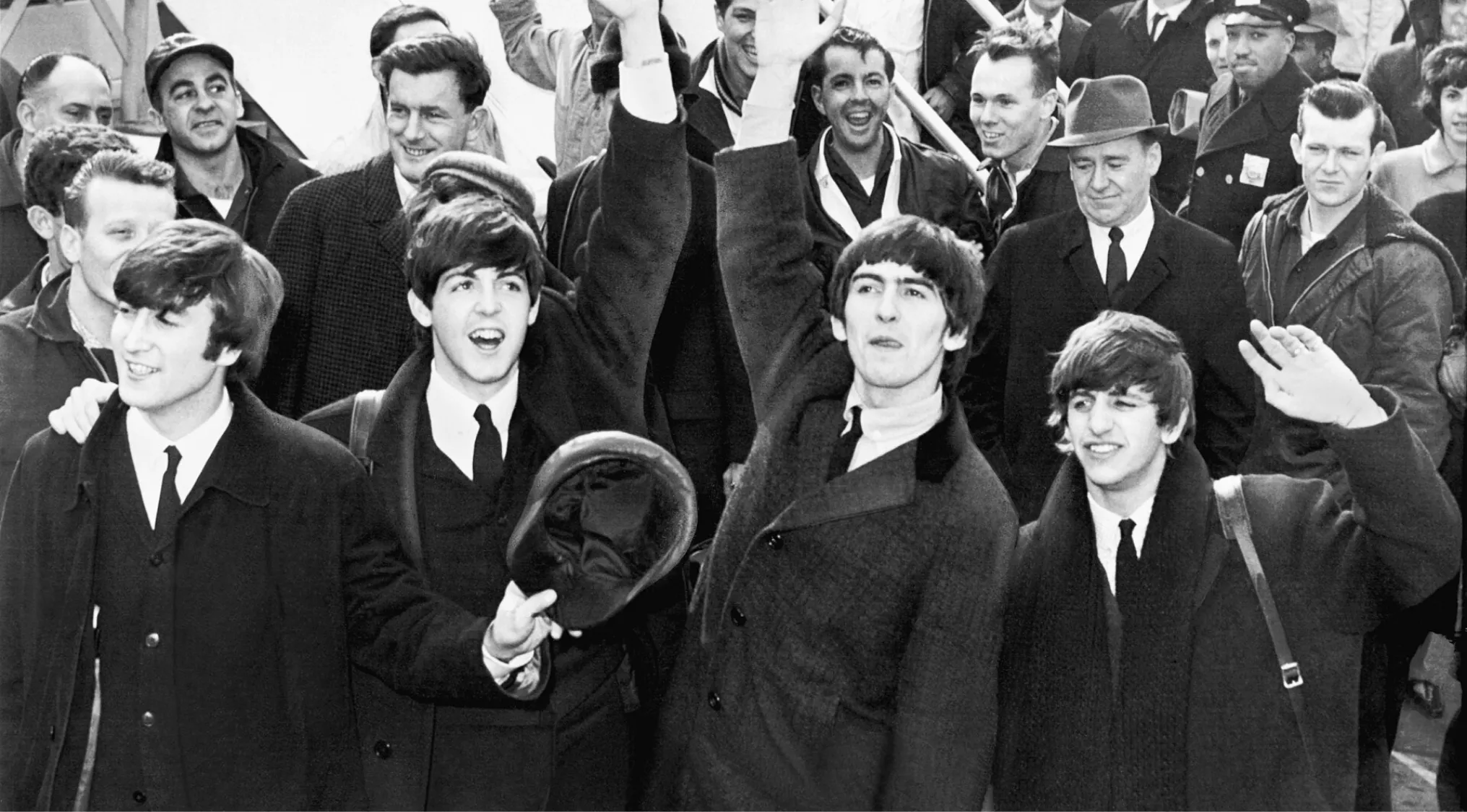 The Beatles waving and smiling to a crowd of fans and onlookers during their early British Invasion in the U.S.