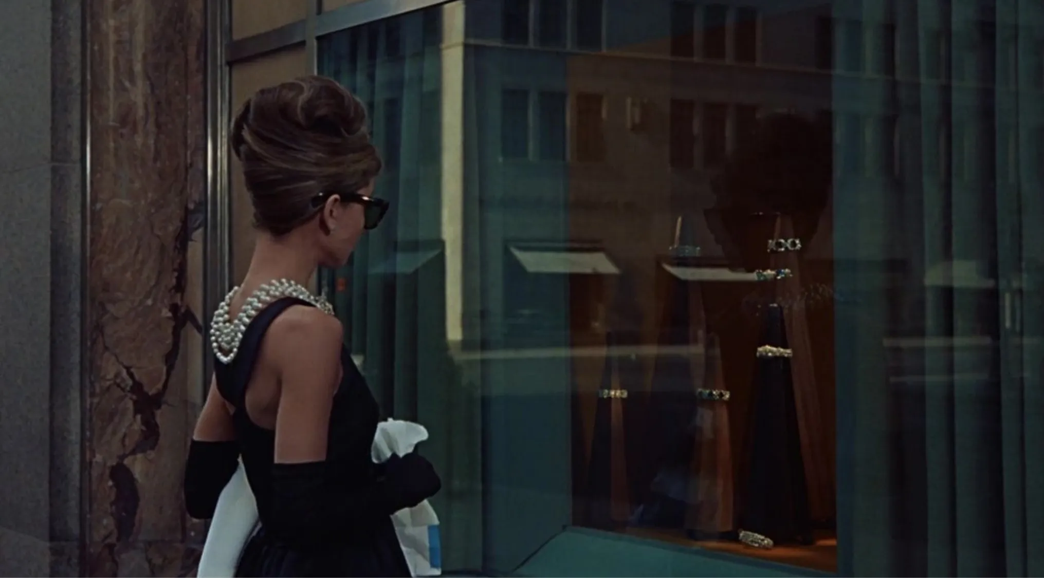 Iconic 60s pop culture scene with a woman in a black dress and pearls looking at a jewelry store window display.