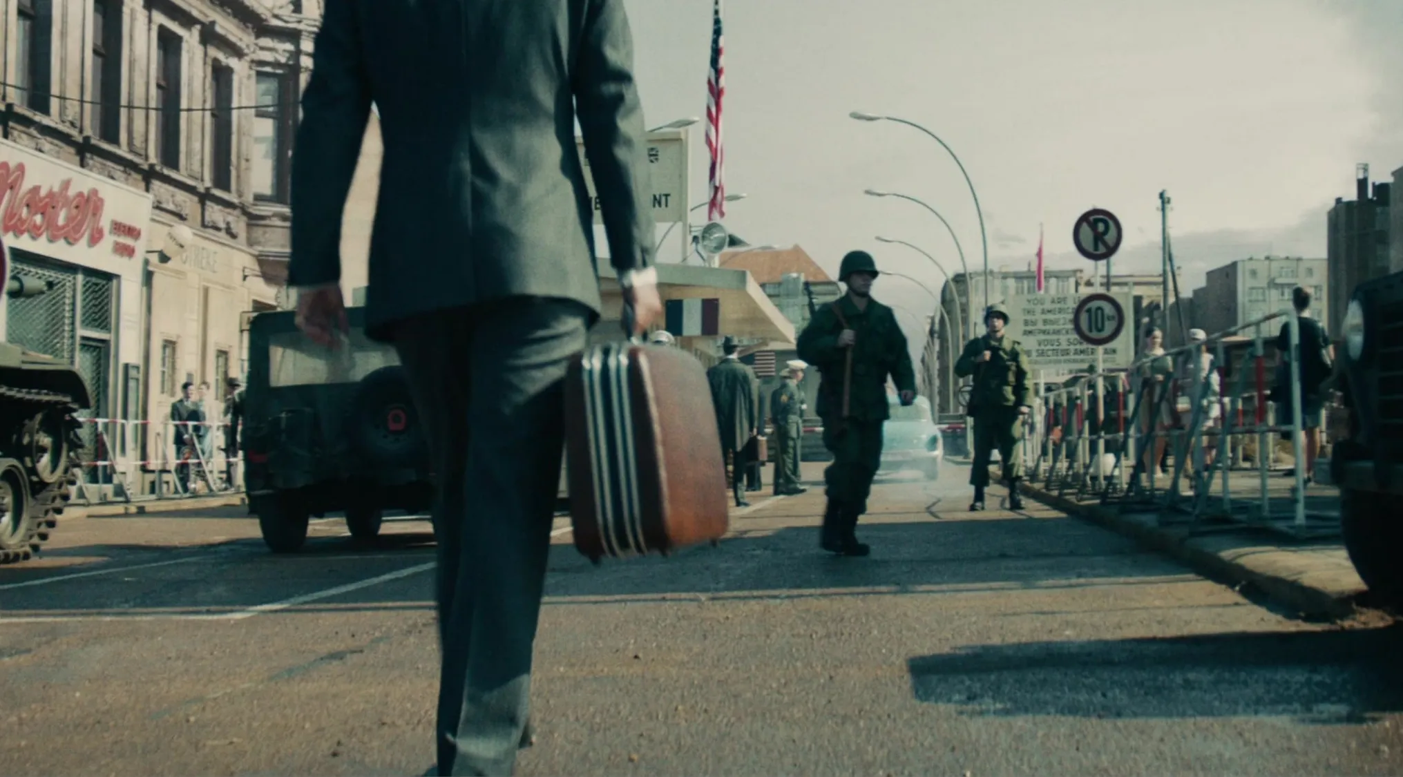 Man in suit carrying briefcase walking through military checkpoint with soldiers and armored vehicles in a spy film series scene.