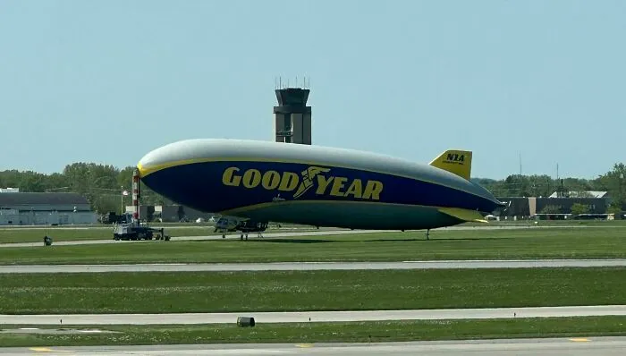 Goodyear blimp parked on an airport runway with control tower and buildings in the background, reflecting made up but real stories.