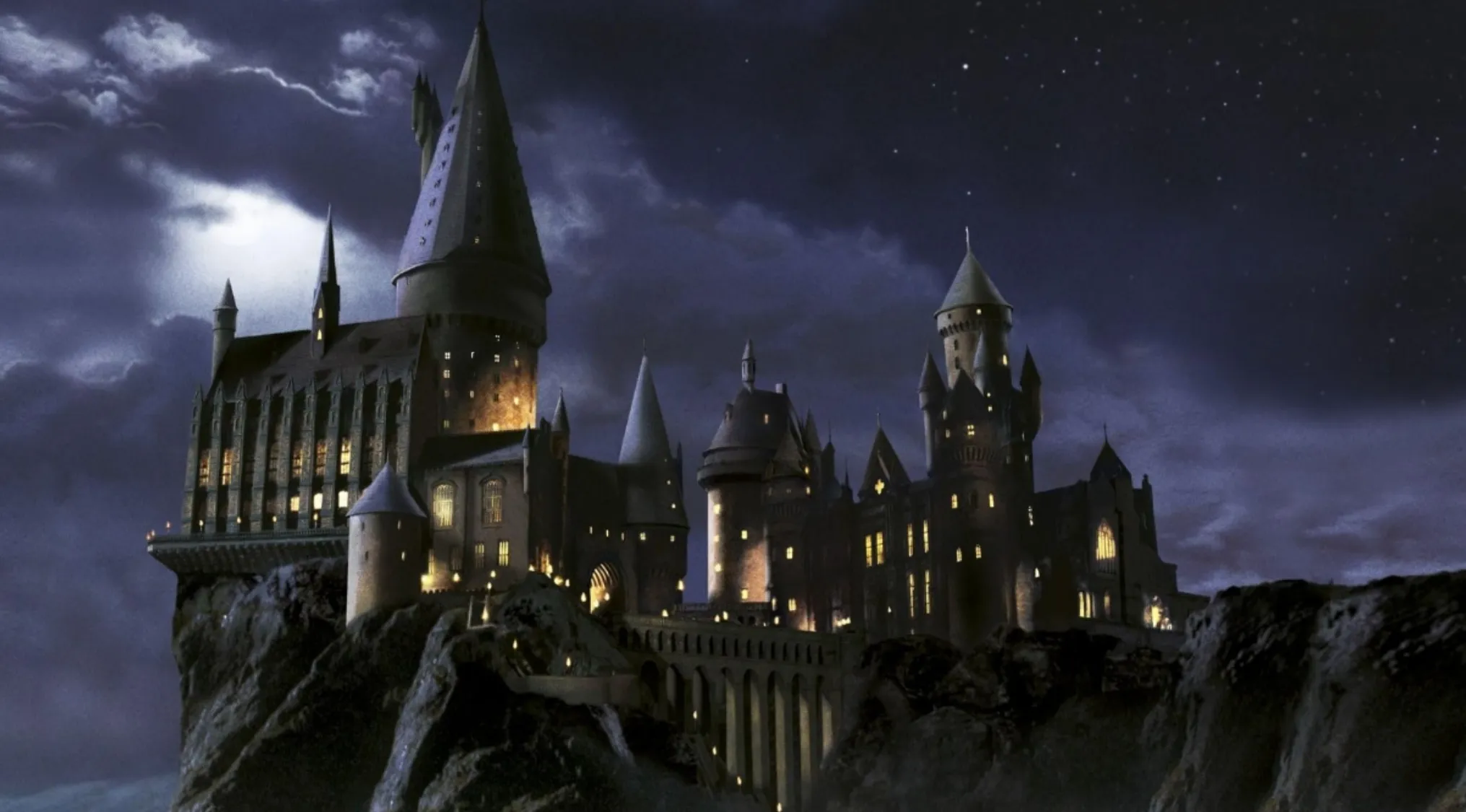 Night view of the wizarding school Hogwarts castle from the Harry Potter franchise with illuminated windows and cloudy sky.