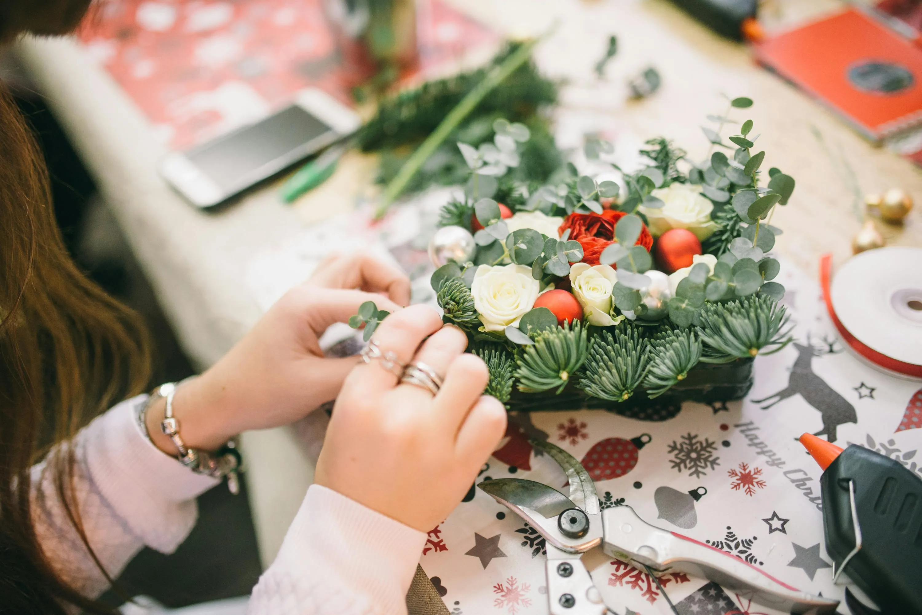 Person working on a DIY project arranging flowers and greenery, surrounded by crafting tools and supplies on a festive table.