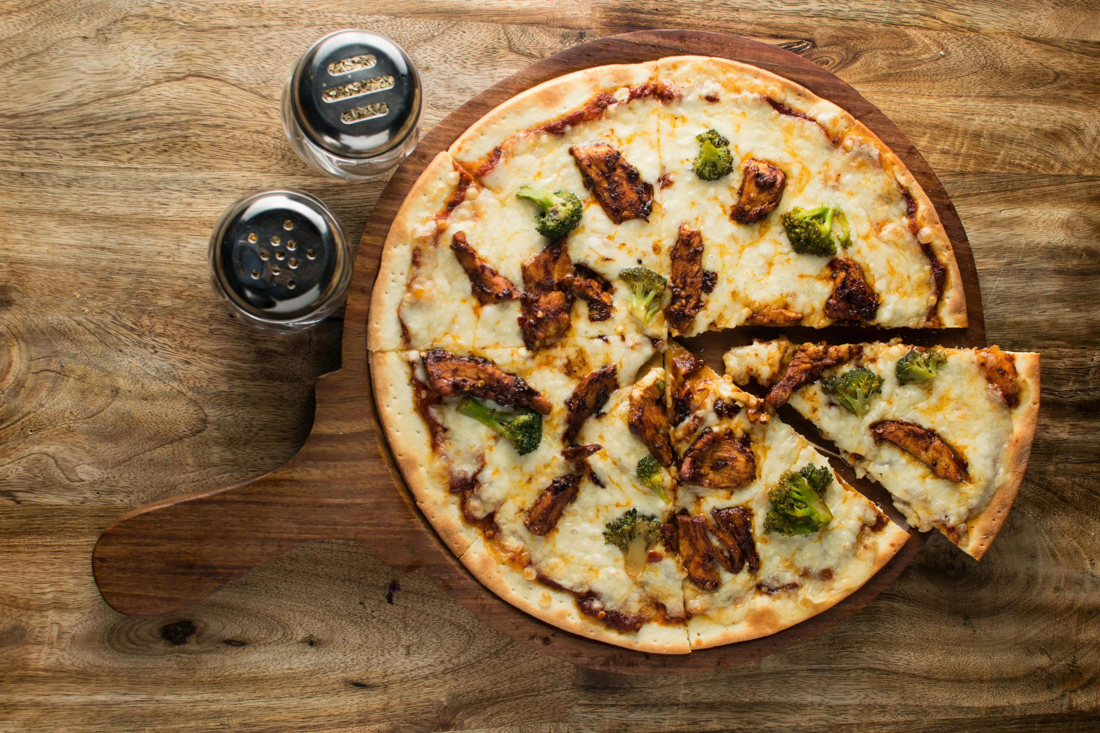 Cafeteria pizza day with a freshly baked pizza topped with chicken and broccoli on a wooden serving board.