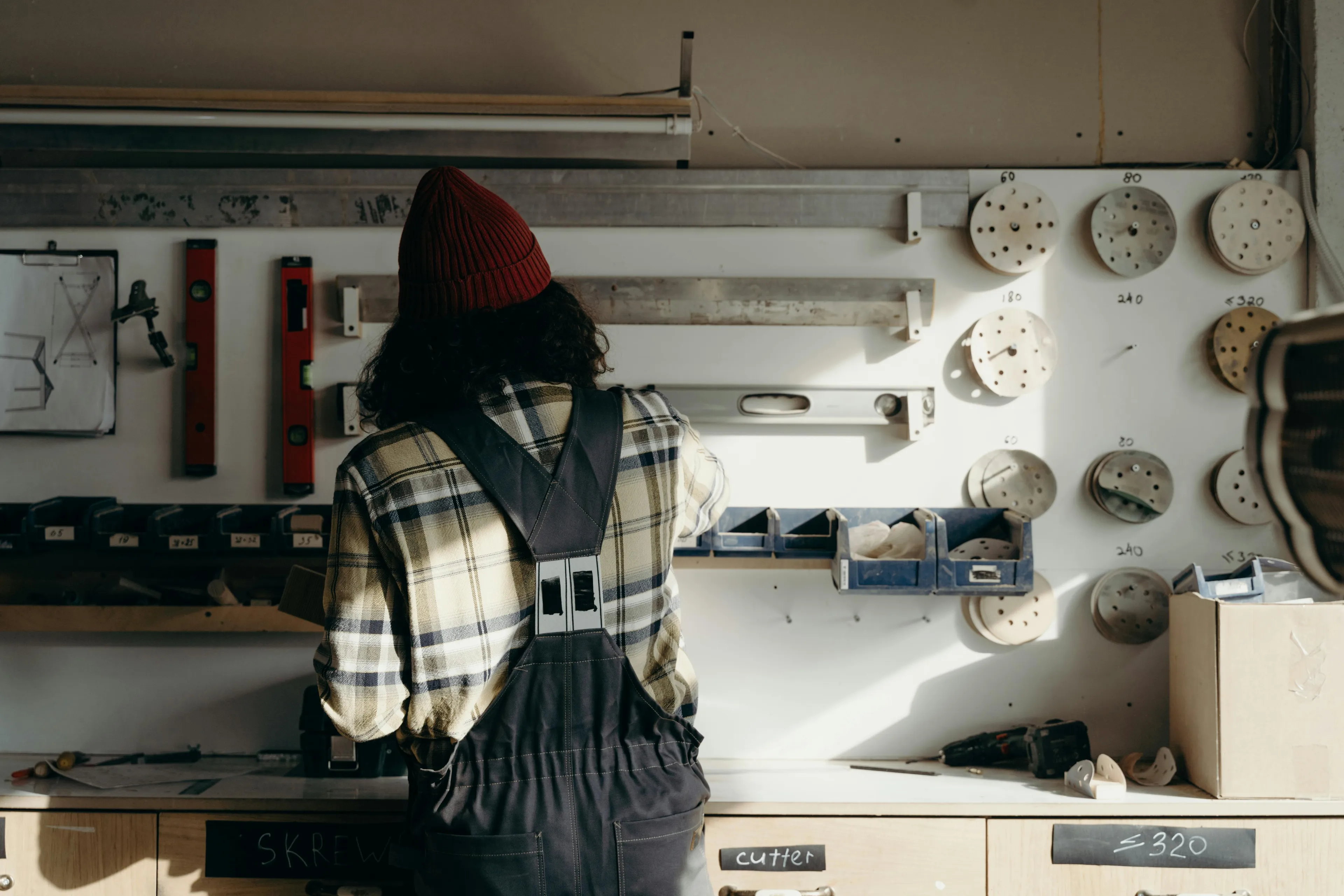 Person in a plaid shirt and beanie working in dad's workshop surrounded by tools and circular wooden pieces on the wall