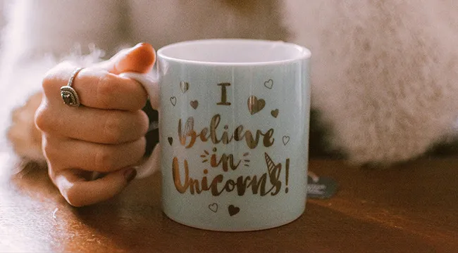 A hand holding a coffee mug with a witty quote saying I believe in unicorns surrounded by small hearts.