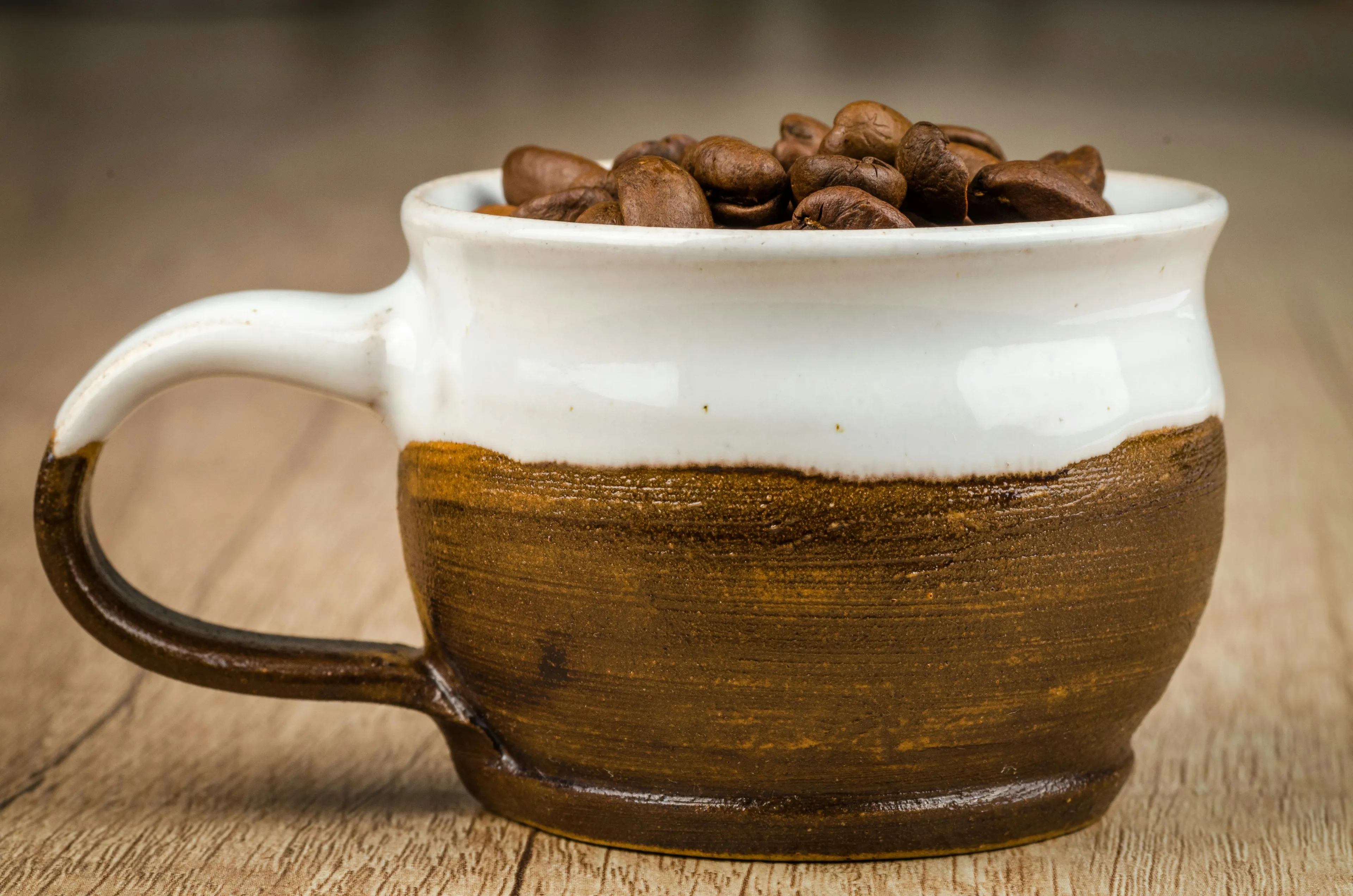 Ceramic coffee mug with a big handle filled with whole coffee beans on a wooden surface.