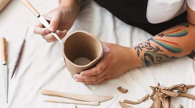 Hands shaping a handmade pottery coffee mug with tools on a table, highlighting a natural and artisanal pottery vibe.