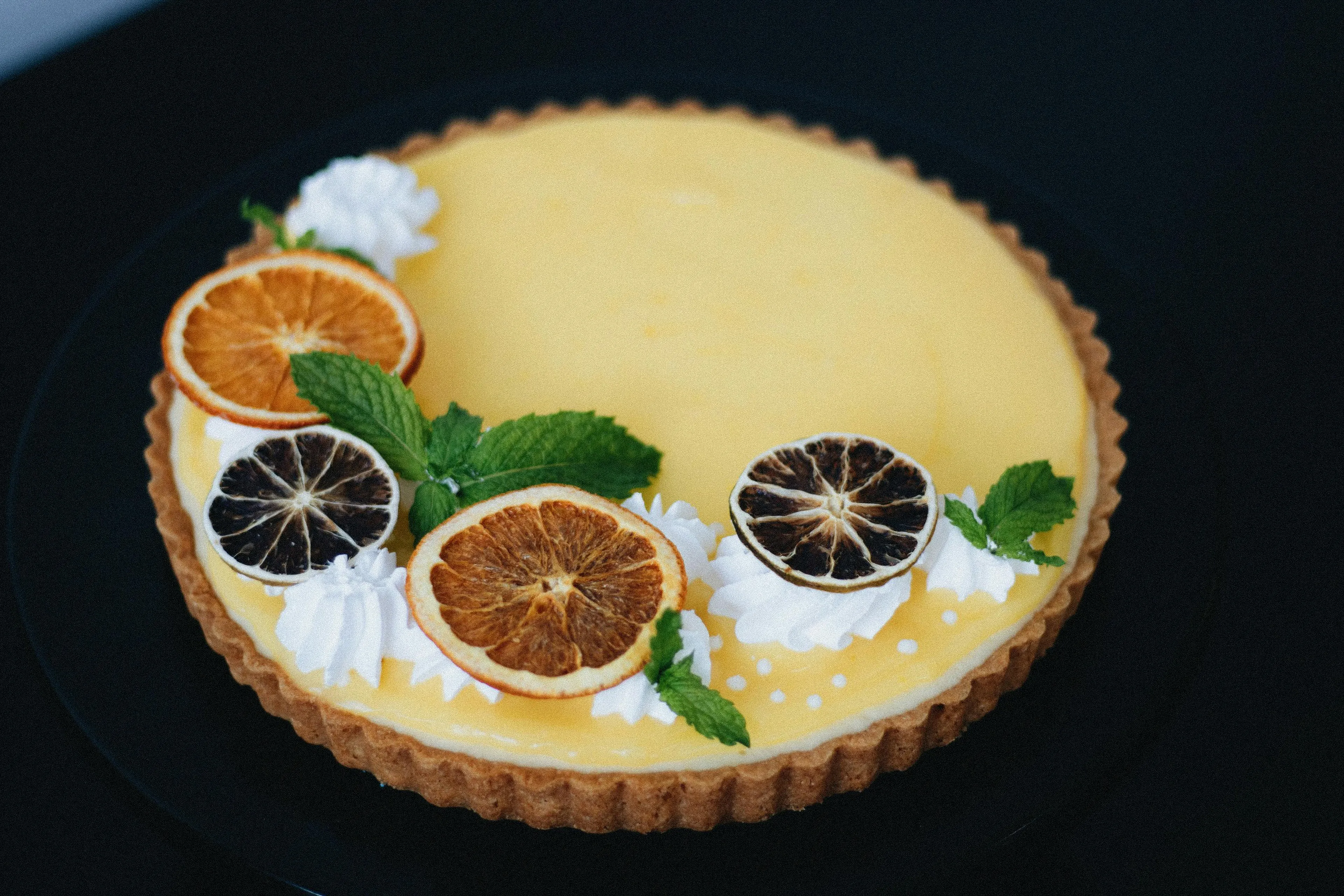 A lemon tart topped with dried citrus slices, whipped cream, and fresh mint leaves on a black plate.