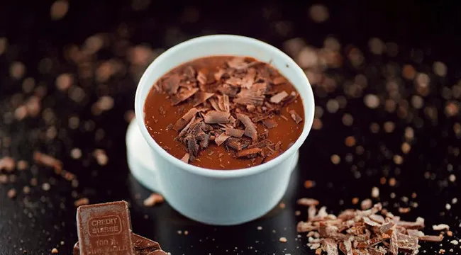 A rich dark chocolate mousse in a white cup garnished with chocolate shavings on a dark surface.