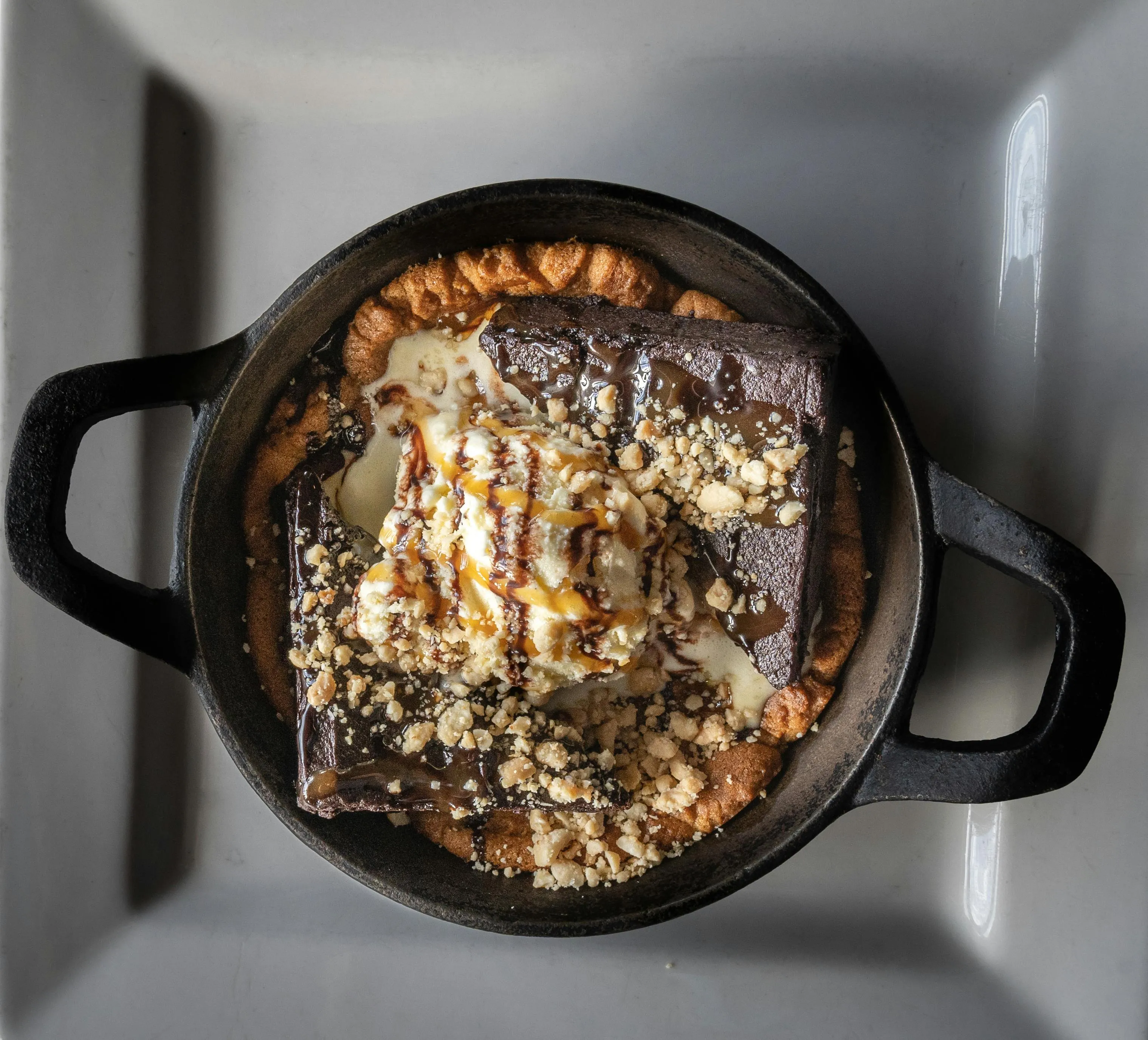 Warm brownie skillet topped with ice cream, caramel drizzle, and crushed nuts served in a cast iron pan.