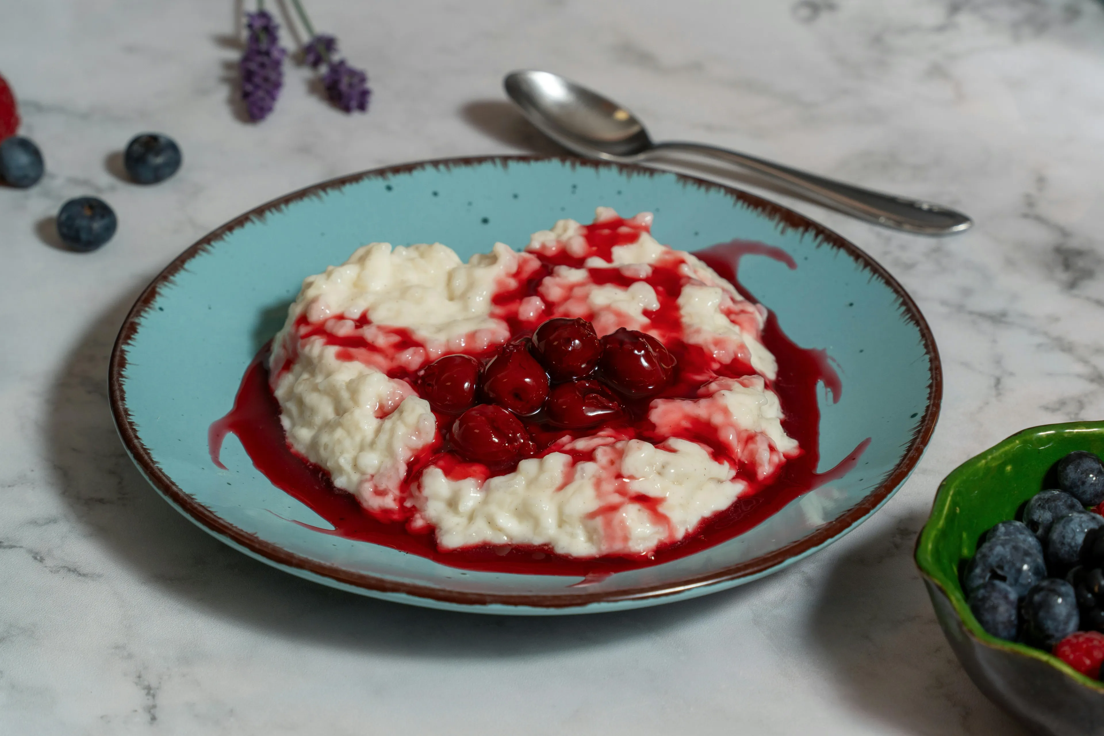 A bowl of creamy rice pudding topped with cherry sauce and whole cherries on a blue plate with a spoon nearby.