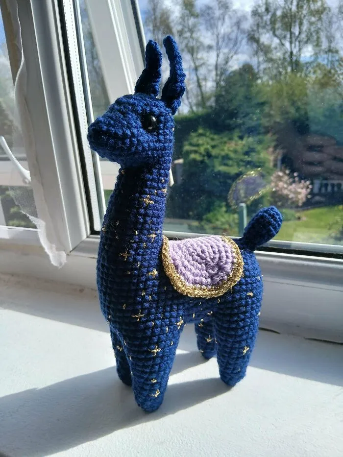 Blue crocheted llama amigurumi with gold accents and purple saddle