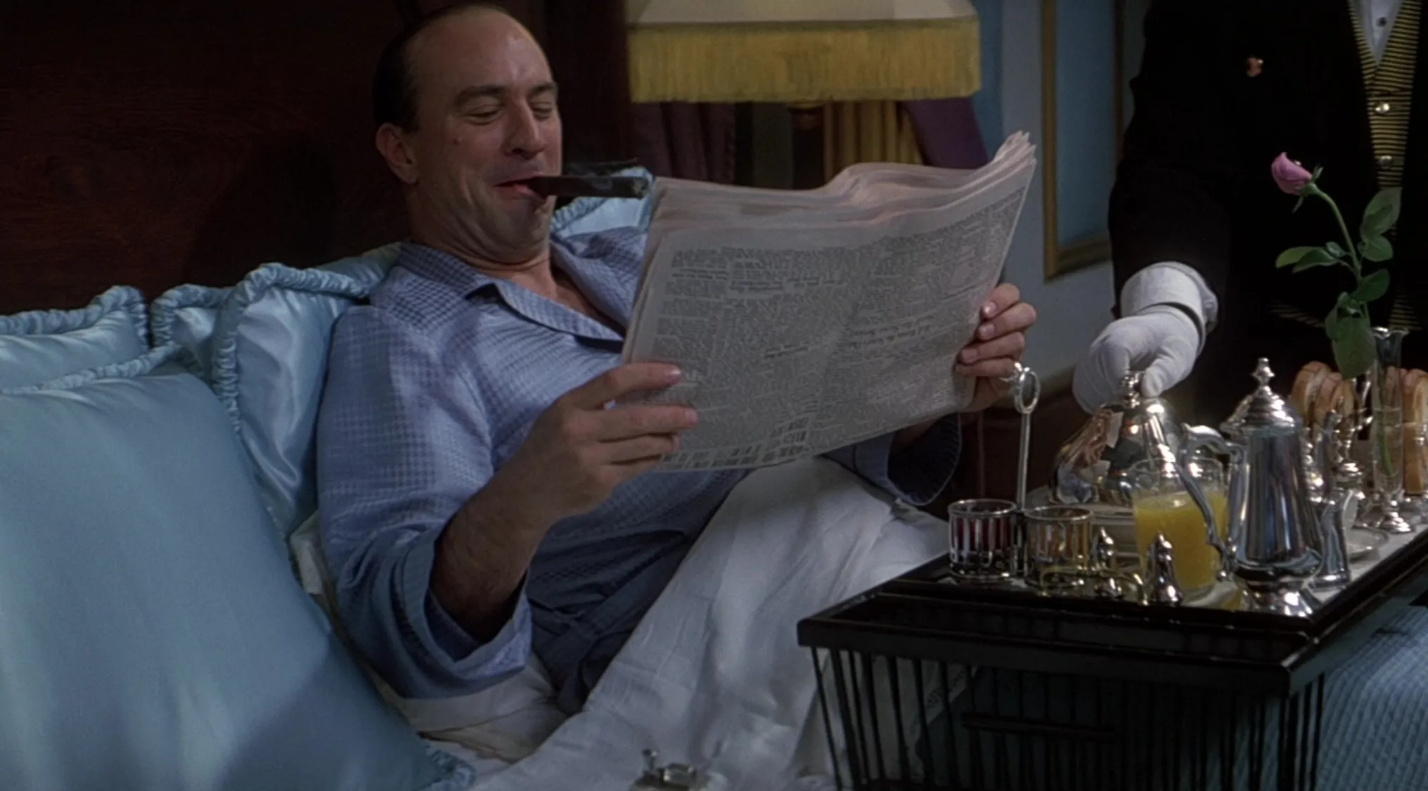 Mob boss in pajamas reading newspaper with cigar, served breakfast in bed by a waiter in a classic movie scene.