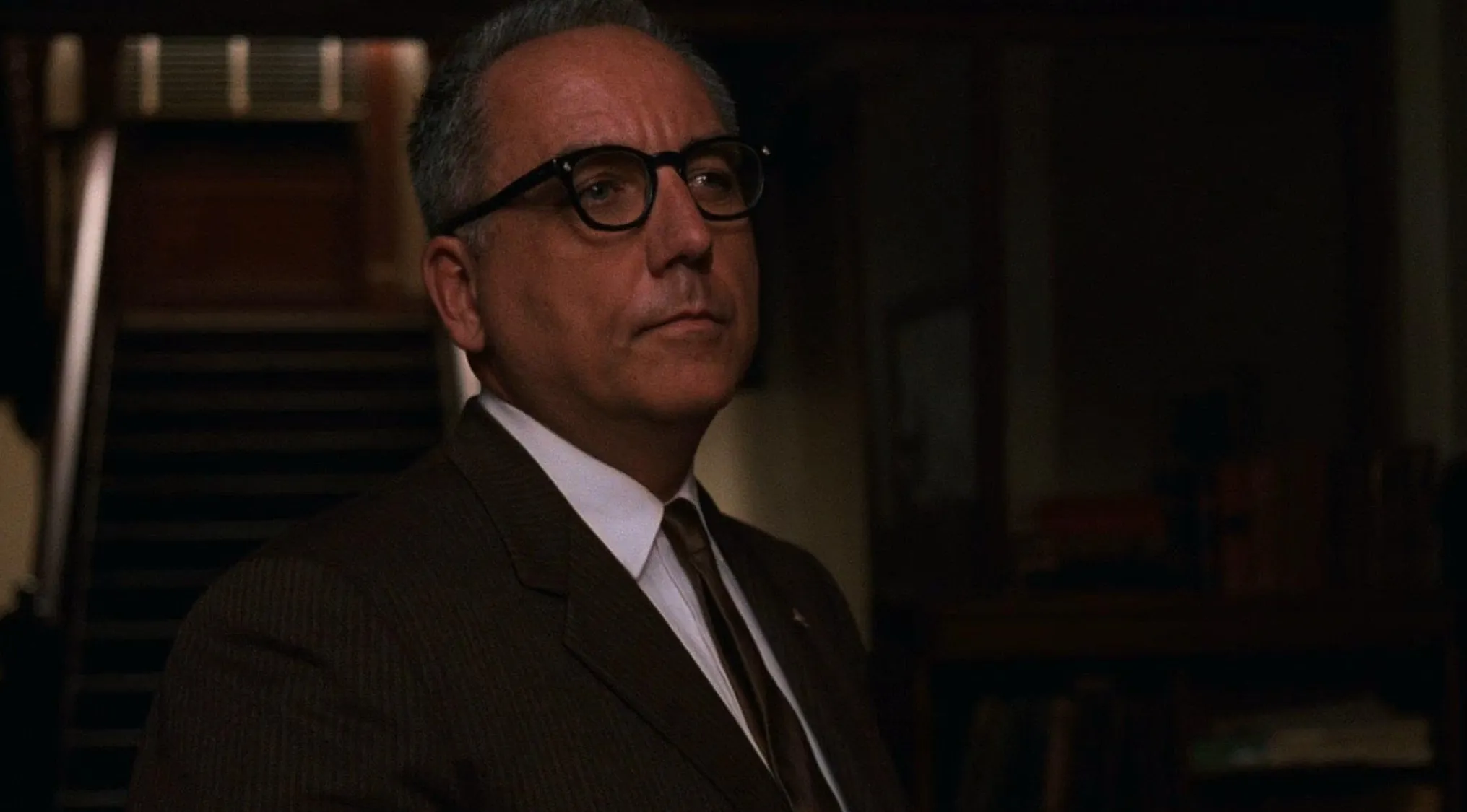 Calm authoritative prison warden wearing glasses and suit, hiding corruption behind religion and discipline.