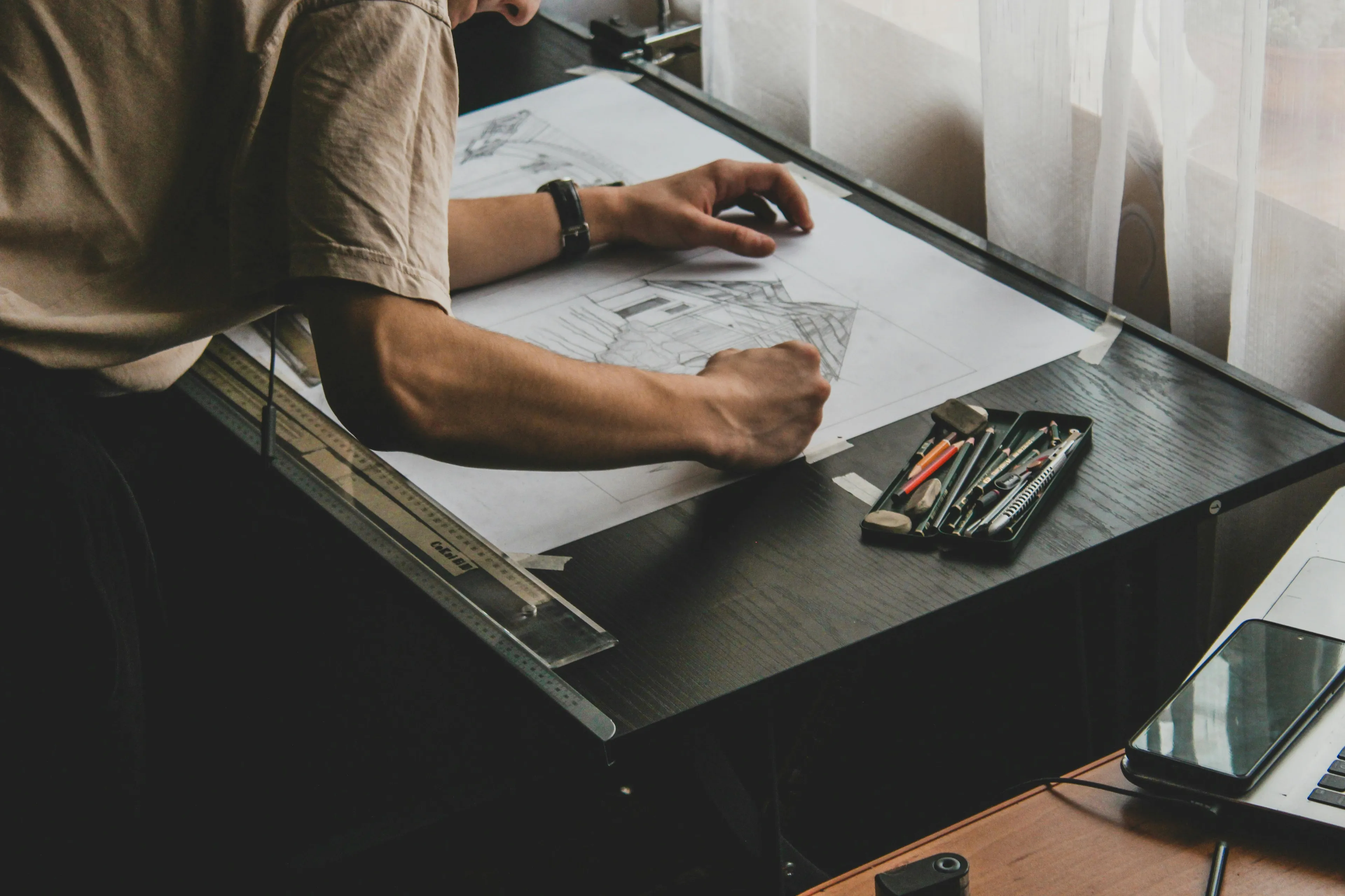 Person using a mid-century drafting table to create architectural drawings with pencils and rulers near a window.