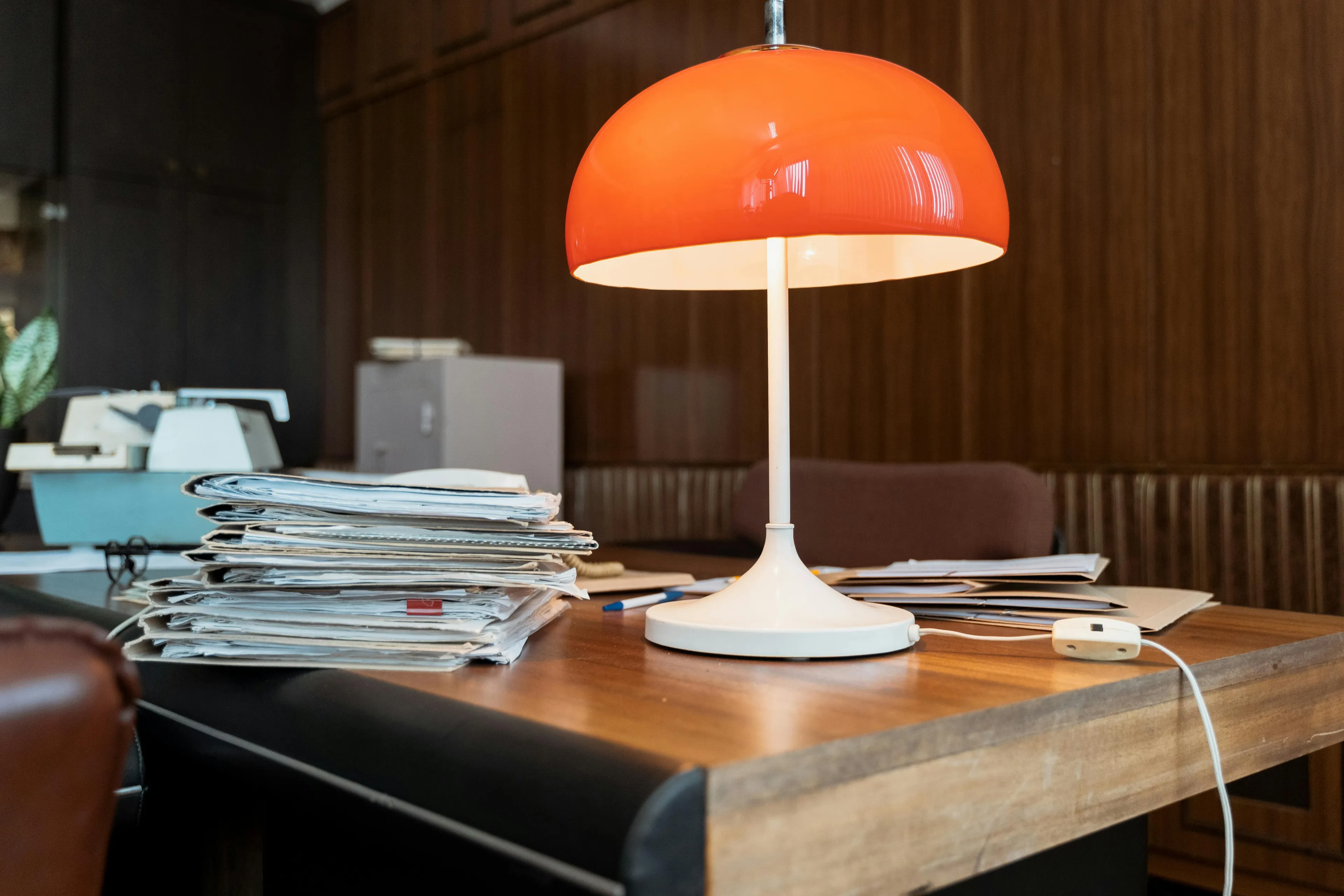 Vintage desk lamp with a rounded orange shade sits on a wooden office desk cluttered with stacked papers.