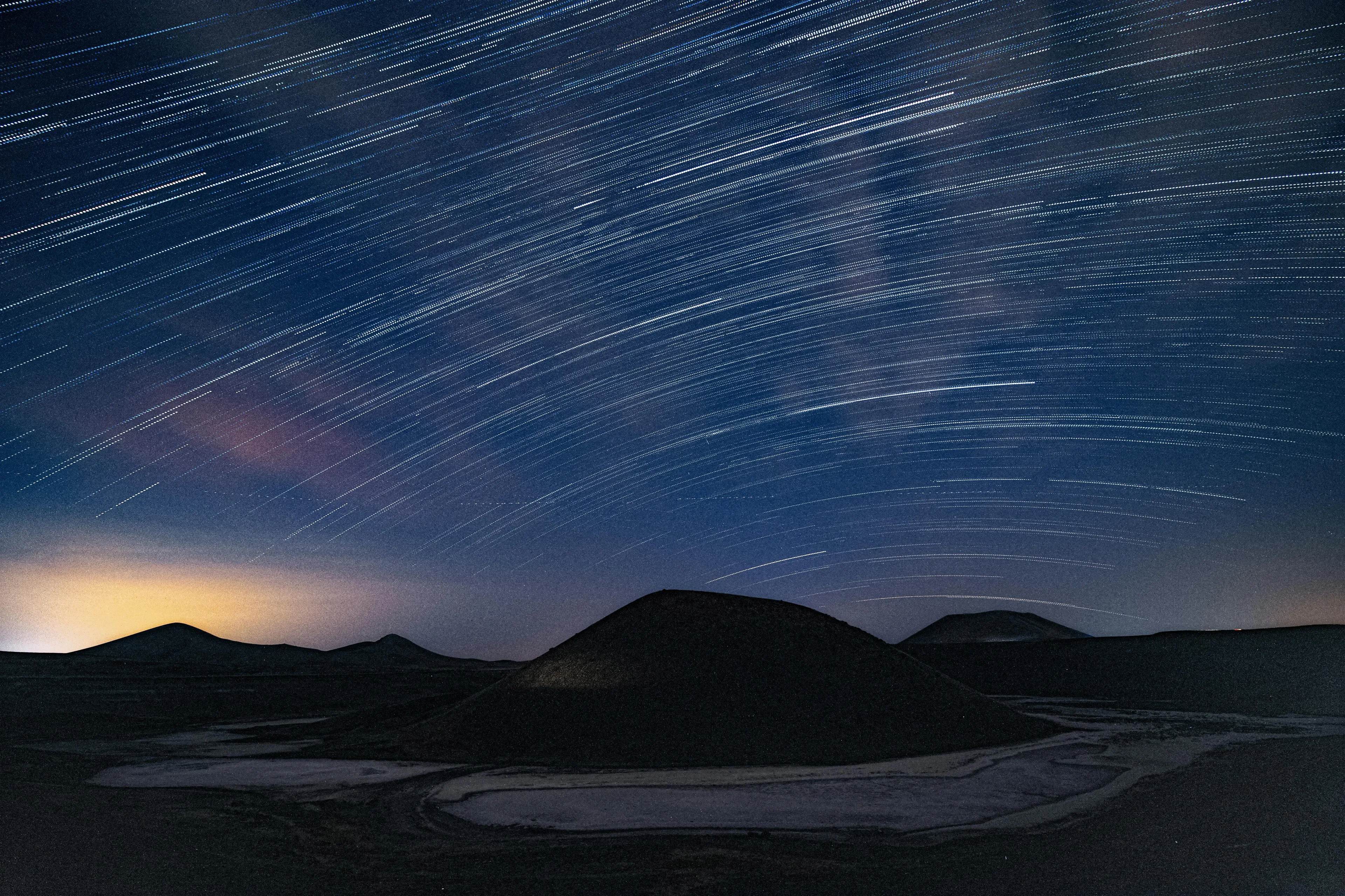 Night sky over dark hills with star trails and meteor shower creating bright streaks across the horizon.