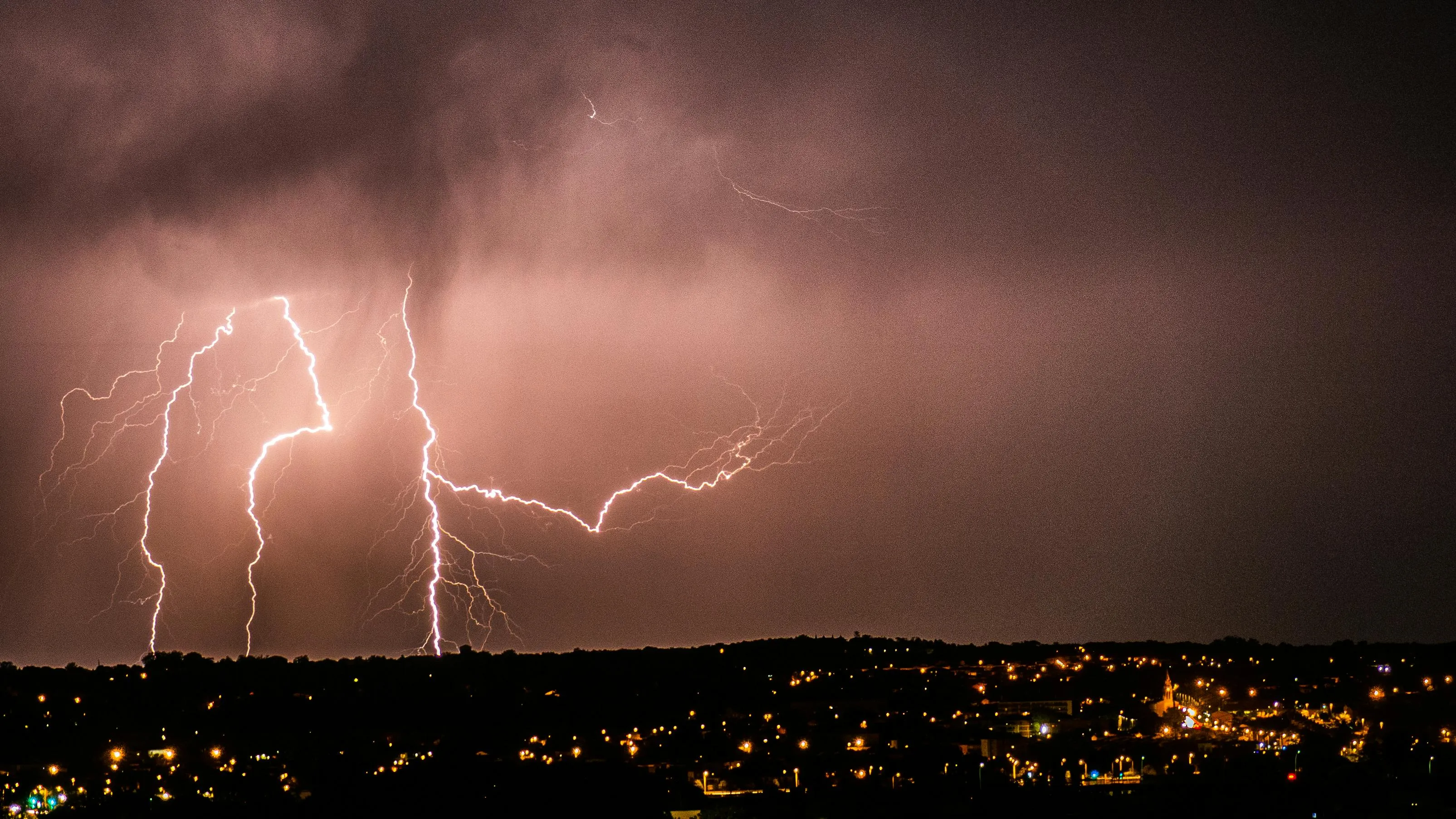 Dramatic thunderstorm with multiple lightning bolts striking over a cityscape at night under dark stormy clouds