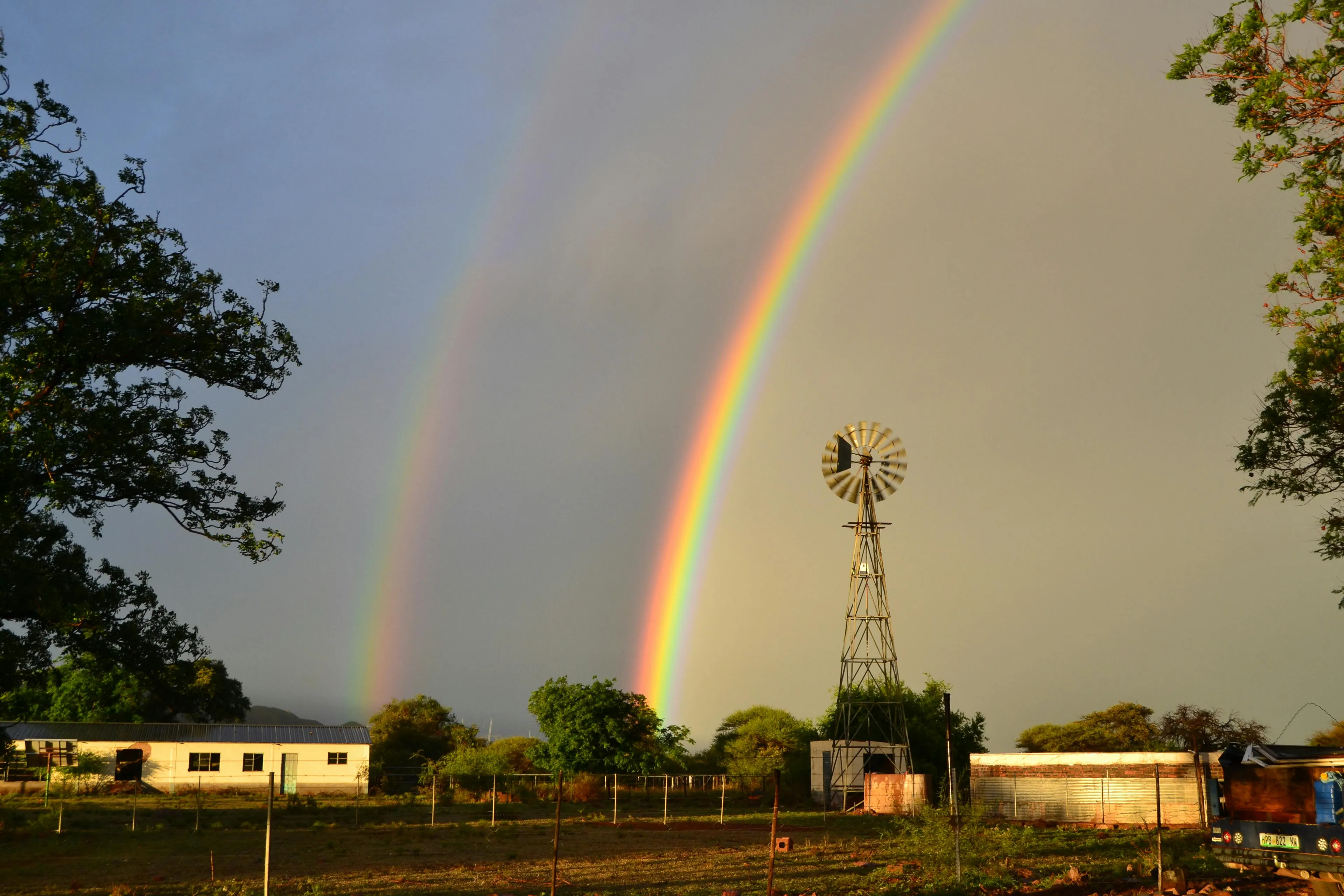 Double rainbow arching over rural landscape with windmill and trees under cloudy sky at sunset.