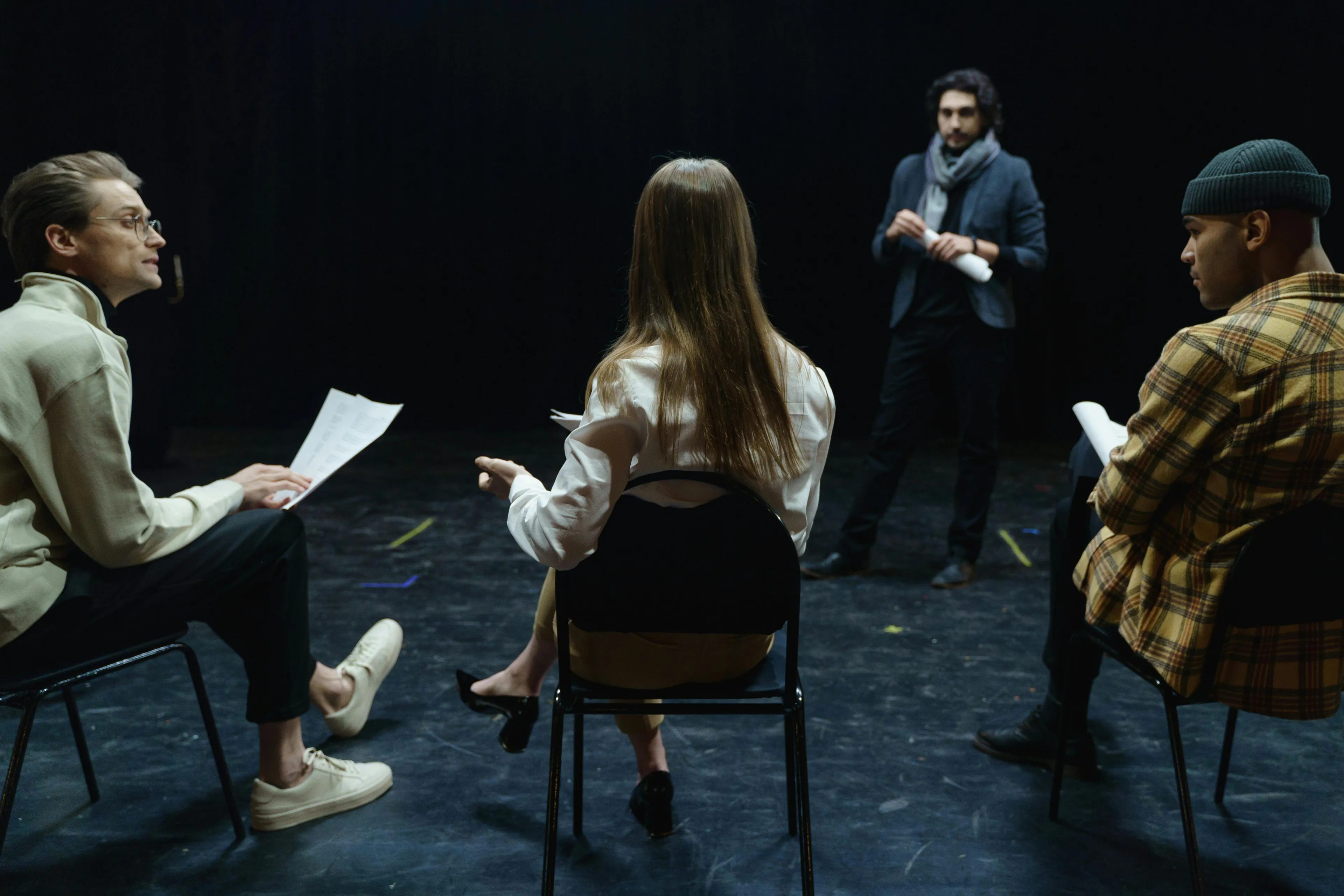 Four adults in a school play monologue rehearsal, seated on chairs, holding scripts and discussing performance.